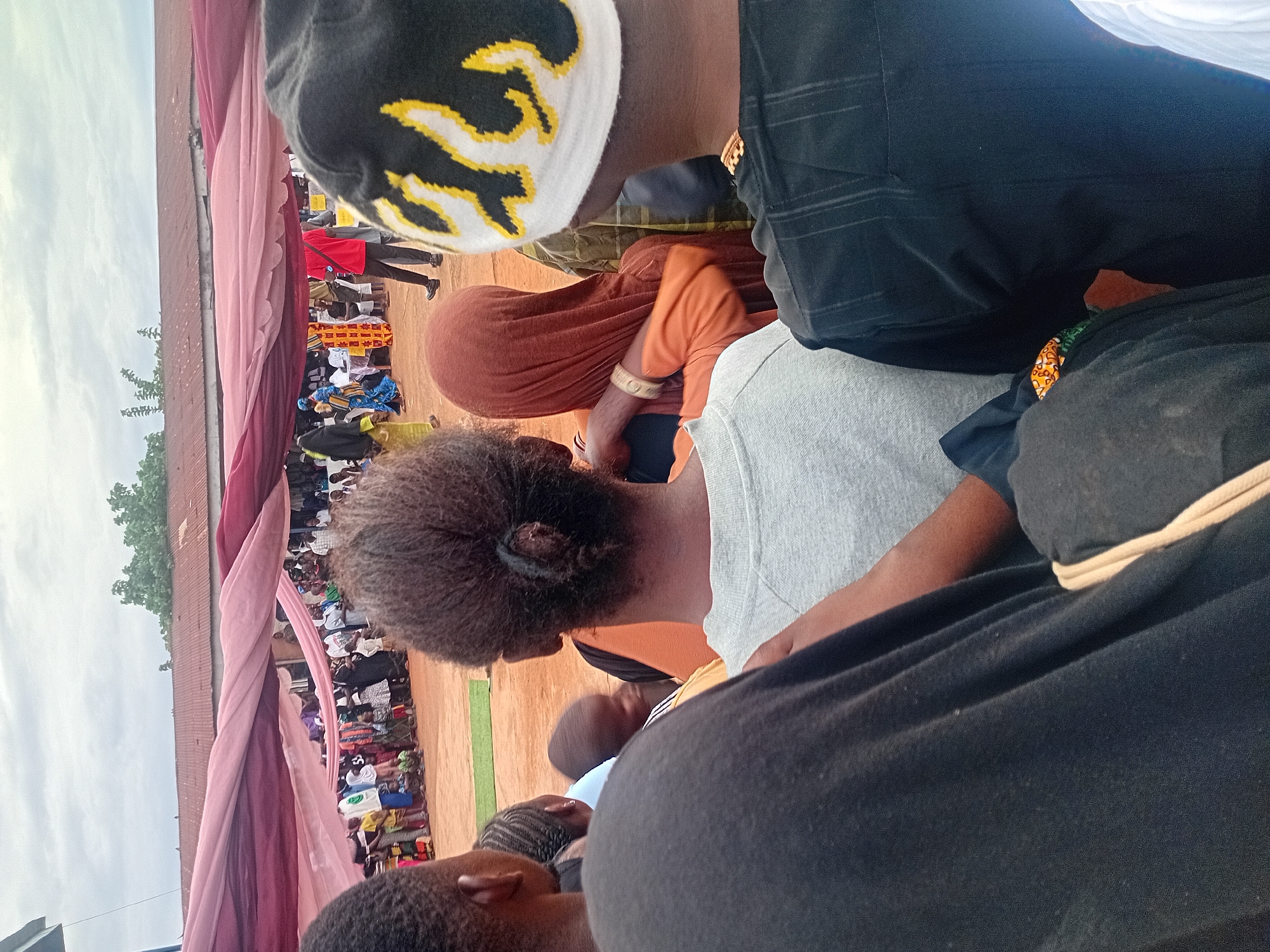 This image depicts an outdoor community gathering in Ogbogbo, Nigeria, under an overcast sky. In the immediate foreground, facing away from the camera, are three individuals. On the left, a person with dark, kinky hair wears a black garment. Centrally, a person with dark hair tied in a bun wears a light grey short-sleeved top, with their right arm extended towards another person wearing a brown headscarf and an orange top, suggesting an interaction or close proximity. On the right, a person wears a black beanie with yellow and white flame-like patterns, a dark striped shirt, and a visible gold chain.
Beyond these foreground figures, a crowd of diverse individuals, including adults and children, is gathered on a reddish-brown dirt ground. A rectangular patch of green turf is visible on the ground in the mid-distance. Overhead, large sheets of draped fabric in shades of pink and burgundy provide temporary shade or decoration. In the mid-ground, a person in a red shirt faces away from the camera, and a woman in a blue patterned dress with a yellow headscarf walks to the left. The background features a building with a reddish-brown corrugated metal roof, with green trees and foliage visible above it. The people in the mid-ground and background are oriented towards a central point, suggesting an ongoing event or performance.