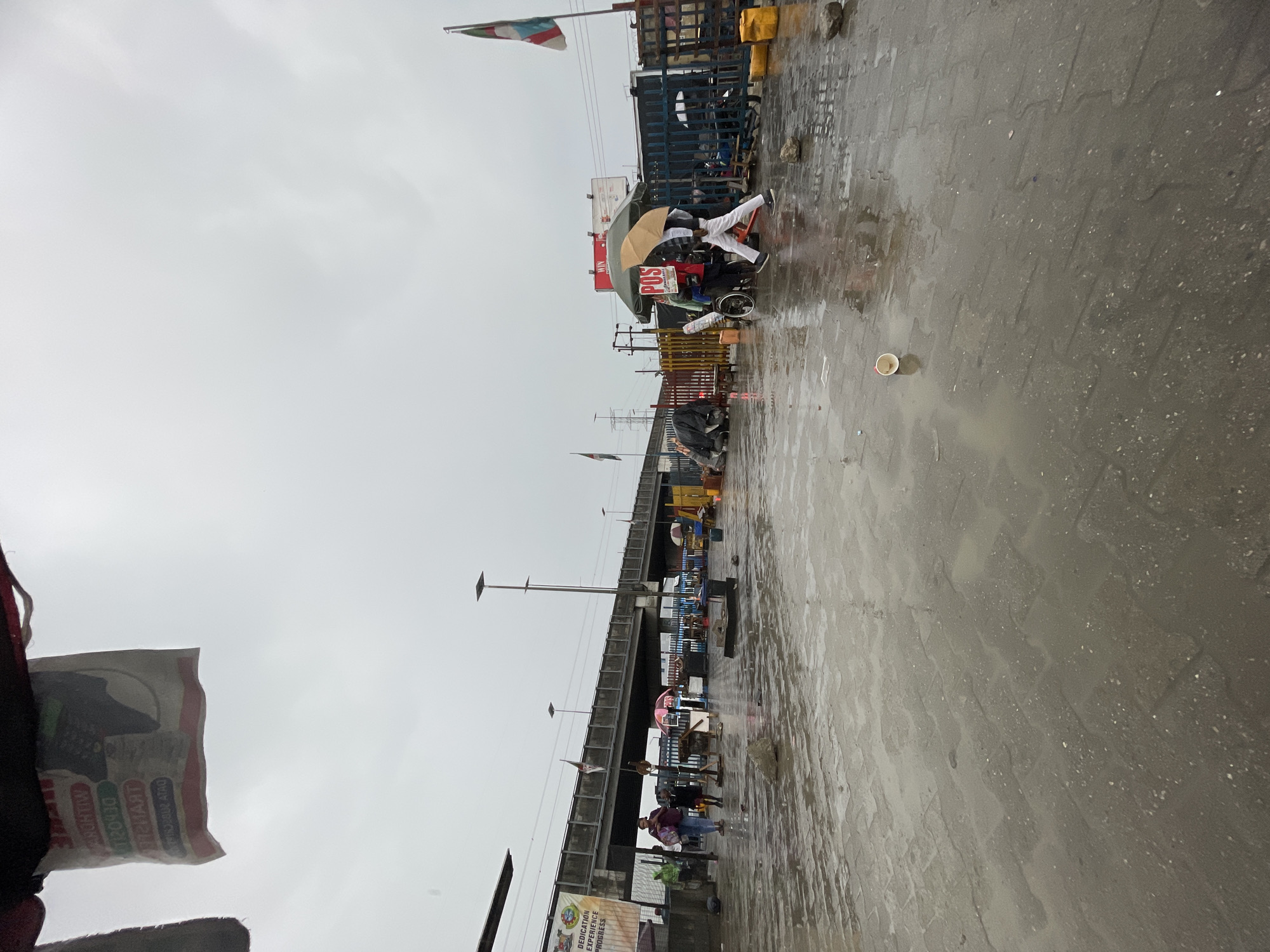 The scene is a street in Lekki, Nigeria on a rainy day. The road is wet and reflects the cloudy sky. A bridge spans overhead. Along the side of the road, there are vendors with stalls and people walking. Some people are using umbrellas. One vendor has a POS system displayed. A flag is visible in the distance. There is a discarded cup on the ground.