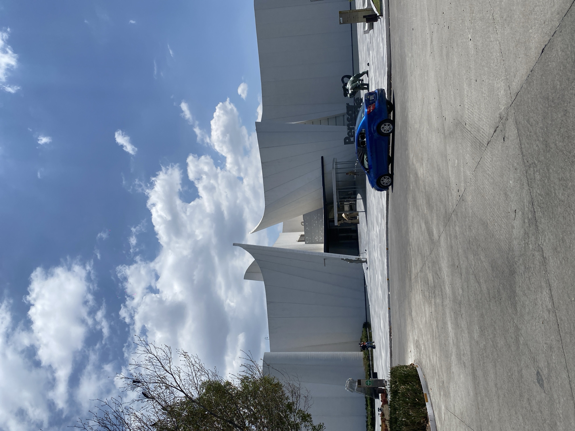 The image shows the Pensacola International Airport terminal in Pensacola, Florida. A blue sedan is parked near the entrance. Sculptures are visible next to the building. The sky is partly cloudy. The surrounding area includes a paved parking area and landscaping. A bare tree is in the lower left corner.