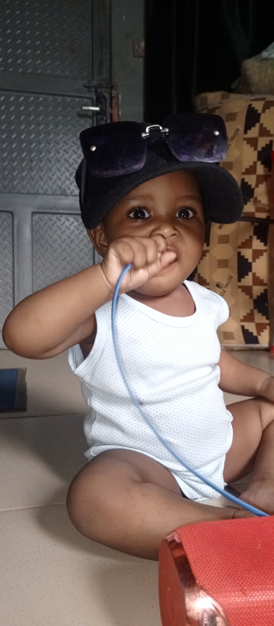 The image depicts an infant seated on the floor, wearing a white sleeveless shirt, a black hat, and oversized sunglasses perched on the hat. The infant has dark skin, and is holding a blue cord in their mouth.
Behind the infant is a metal door, and patterned boxes. A portion of a red object is visible in the foreground.
The location in the city of Keffi, Nigeria is unknown.
