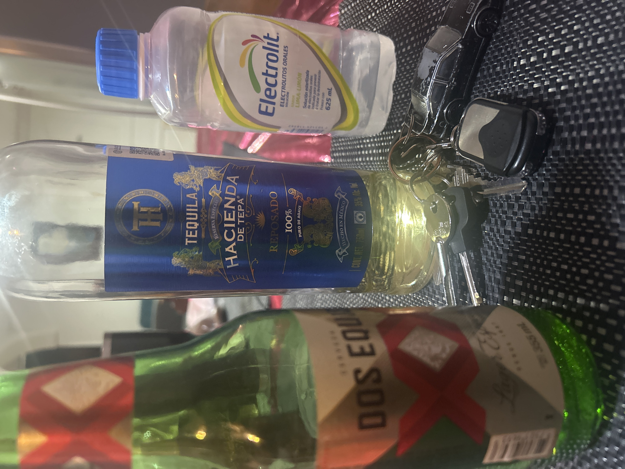 A tabletop setting with several items is shown. The dominant objects in the view, oriented horizontally from bottom to top, include a green bottle of Dos Equis beer, a bottle of Hacienda de Tepa Tequila, a bottle of Electrolit, and a set of car keys. The bottles appear to be nearly empty. The table has a textured black and grey woven surface. The scene appears to be indoors. It is not possible to determine if it is located in the city of Puebla, Mexico.