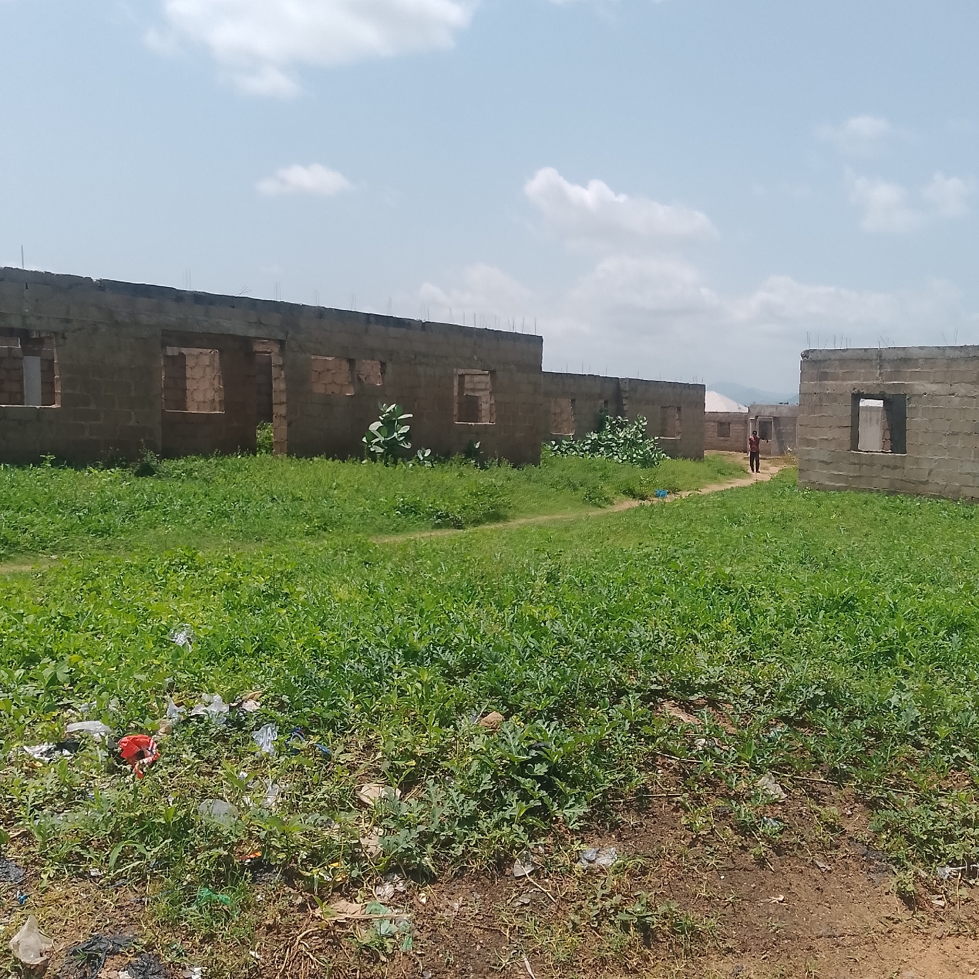 This is an outdoor, eye-level, medium shot of an unfinished construction site in Jalingo, Nigeria. The foreground is dominated by lush green grass and scattered debris, including plastic bags and a red object. In the midground, a person in a red shirt and dark pants stands on a dirt path between two partially constructed brick buildings. These buildings appear to be homes or commercial spaces, with open doorways and window frames visible. The walls are made of concrete blocks. In the background, more unfinished structures and a distant hillside are visible under a bright, cloudy sky. The overall impression is one of ongoing construction and an undeveloped area.