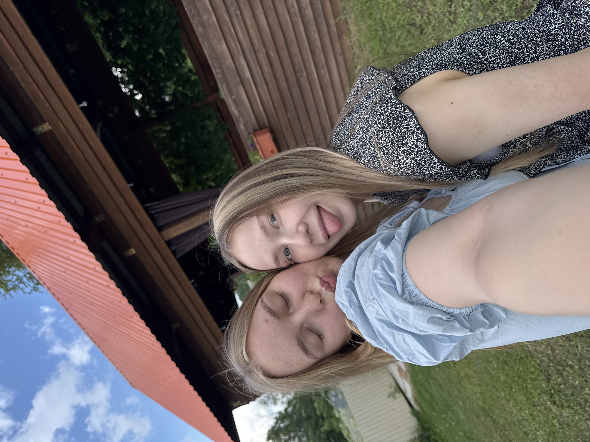 Two young women are taking a selfie outdoors, in front of a wooden structure with a red corrugated roof. The woman on the left is wearing a light blue dress and is puckering her lips for a kiss. The woman on the right is wearing a black and white patterned dress and is sticking her tongue out. Both women have blonde hair. The background shows green grass, trees, and a cloudy blue sky. This scene is consistent with the location of Zhigalki, Russia, where such outdoor recreational areas are common.