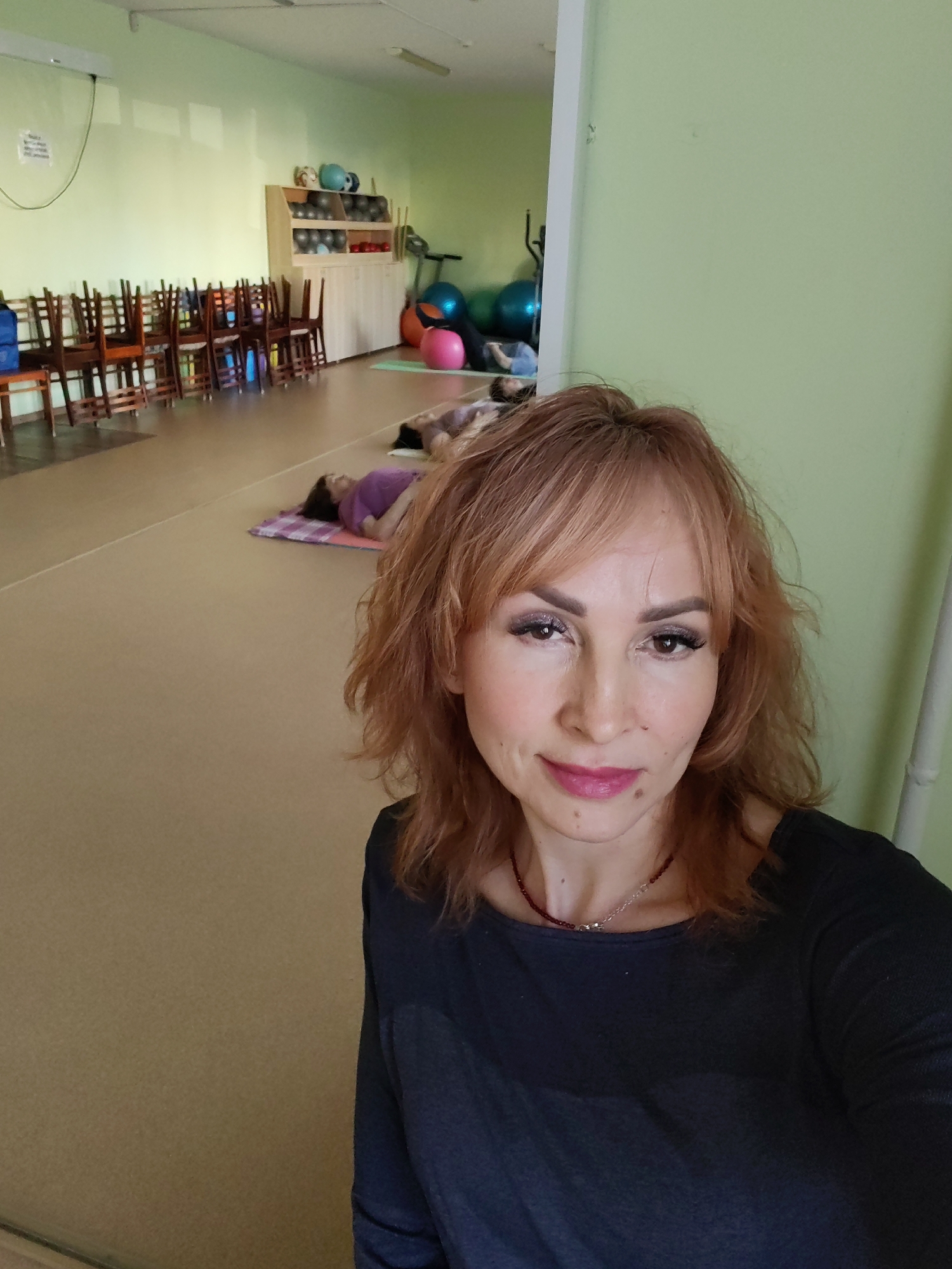 The image shows a woman taking a selfie in what appears to be a fitness or yoga studio. In the background, several people are lying on mats on the floor, possibly engaged in a relaxation exercise. There are exercise balls, a treadmill, a storage unit filled with equipment, and a row of stacked chairs. The walls are pale green, and the floor is beige. The image's origin is Chaikovsky, Russia, based on information available from sources related to the original image.