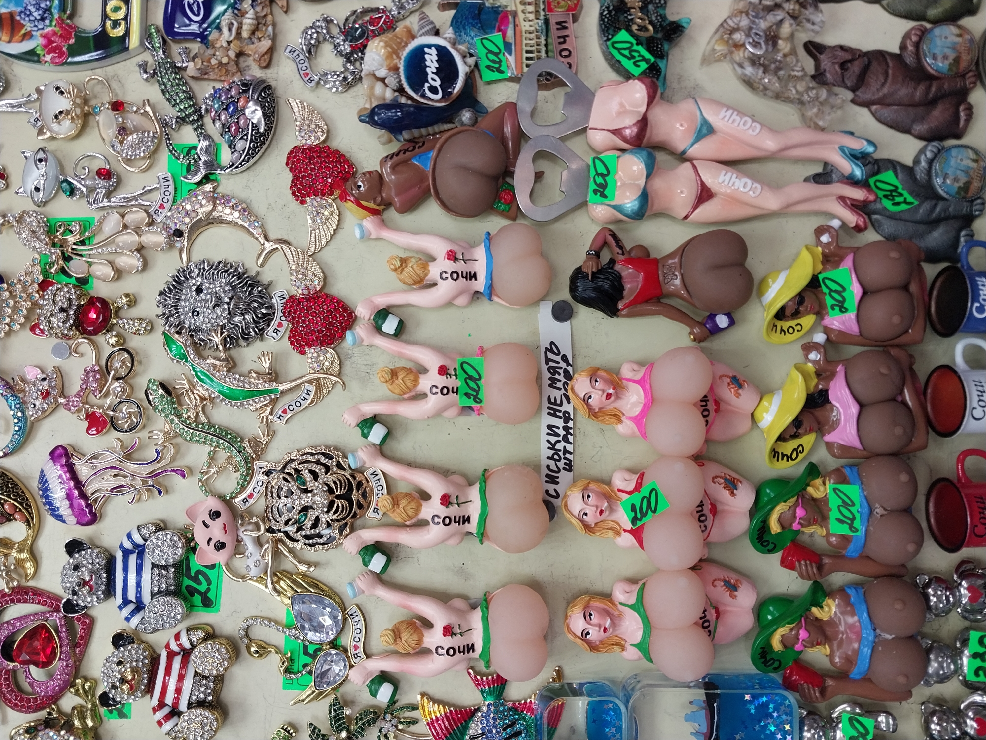 This overhead shot captures a dense display of various souvenirs laid out on a light-colored, flat surface, suggesting a market stall or shop. The items are tightly packed, showcasing a wide range of designs and materials.
A significant portion of the display features stylized human figures, predominantly female, made of plastic or resin. These figures often have exaggerated buttocks and breasts, and many are depicted in bikinis or skimpy clothing. A common design integrates these figures with metal bottle openers, serving as both novelties and functional items. Many of these figures bear the Russian word "СОЧИ" (Sochi), often written on the buttocks or clothing, identifying them as tourist merchandise from the city of Sochi.
Interspersed among the figures are numerous decorative brooches, pins, or magnets made of metal and adorned with gemstones or rhinestones. These include designs such as lizards, dolphins, jellyfish, cats, swans, hearts, abstract floral patterns, and other animal shapes like bears and pandas. Some items are also shaped like miniature cars.
Small green price tags are visible on many items, indicating prices such as "200," "250," and "280," likely in Russian Rubles. A prominent detail is a piece of paper or cardboard near the suggestive figures, displaying the Russian text "СИСЬКИ НЕ МЯТЬ ШТРАФ 200," which translates to "DO NOT SQUEEZE BREASTS, FINE 200."
The items display a diverse color palette, including metallic silvers and golds, vibrant reds, blues, greens, and various skin tones. The overall impression is of a bustling and eclectic souvenir stand aimed at tourists. No people are visible in the image; the focus is entirely on the merchandise.