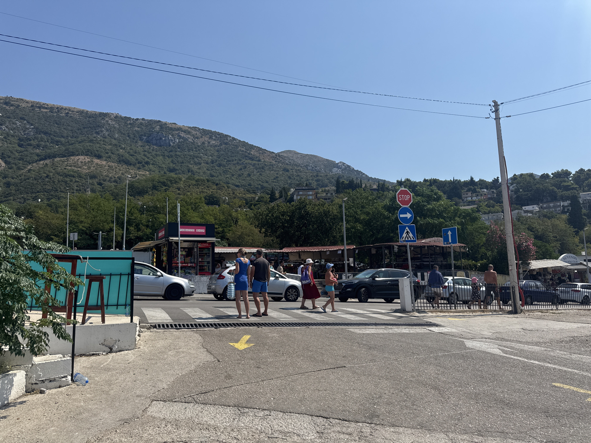 The image depicts a street scene in Sutomore, Montenegro, on a clear, sunny day. In the foreground, a pedestrian crossing is visible, marked with white stripes and a yellow directional arrow on the asphalt. Several people are crossing the street, including a group of four individuals on the left, two men and two women, and a single person further right, a woman with a red shawl. A black SUV is stopped at the crossing, and a silver car is also present. To the left of the crossing, a turquoise structure with stools and a fence is visible, with a plastic bottle on the ground in front of it. In the background, there are several small shops and kiosks, one with "TOBACCO SHOP" signage. Beyond the shops, lush green trees and vegetation cover the lower slopes of a large, rocky mountain that dominates the landscape under a bright blue sky. A utility pole with wires stands on the right side of the image, with traffic signs including a "STOP" sign and pedestrian crossing signs. The overall impression is a lively coastal town environment.