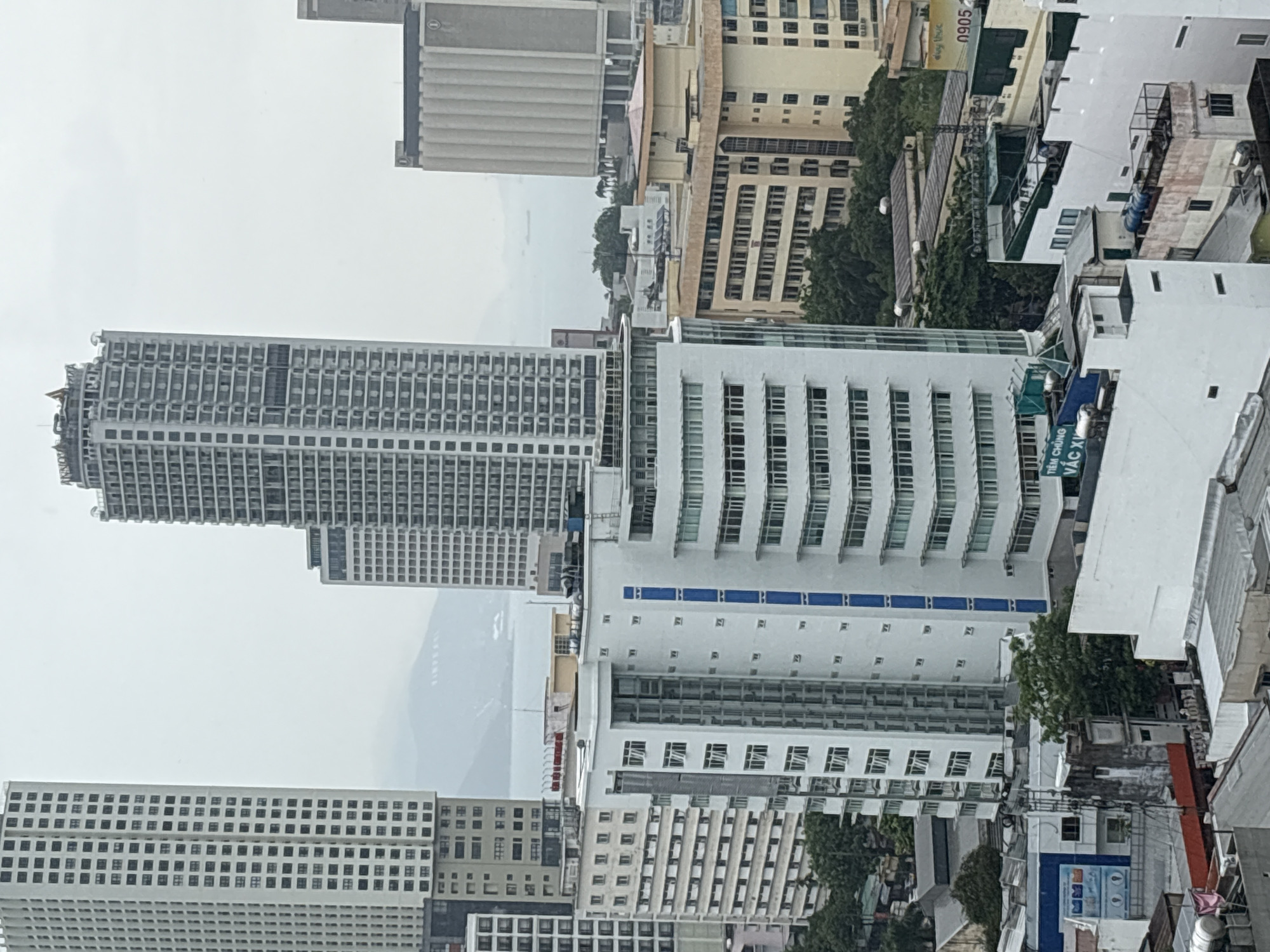 The image shows a cityscape in Nha Trang Ward, Vietnam, featuring several tall buildings against a cloudy sky. Predominantly, there are high-rise structures with grid-like window patterns and white or light-colored facades. Some buildings have blue accent stripes. There is a partial view of mountains in the distance, obscured by haze. The cityscape includes residential and commercial buildings, with some greenery intermixed amongst the urban structures. The built environment dominates, indicating a dense urban setting.