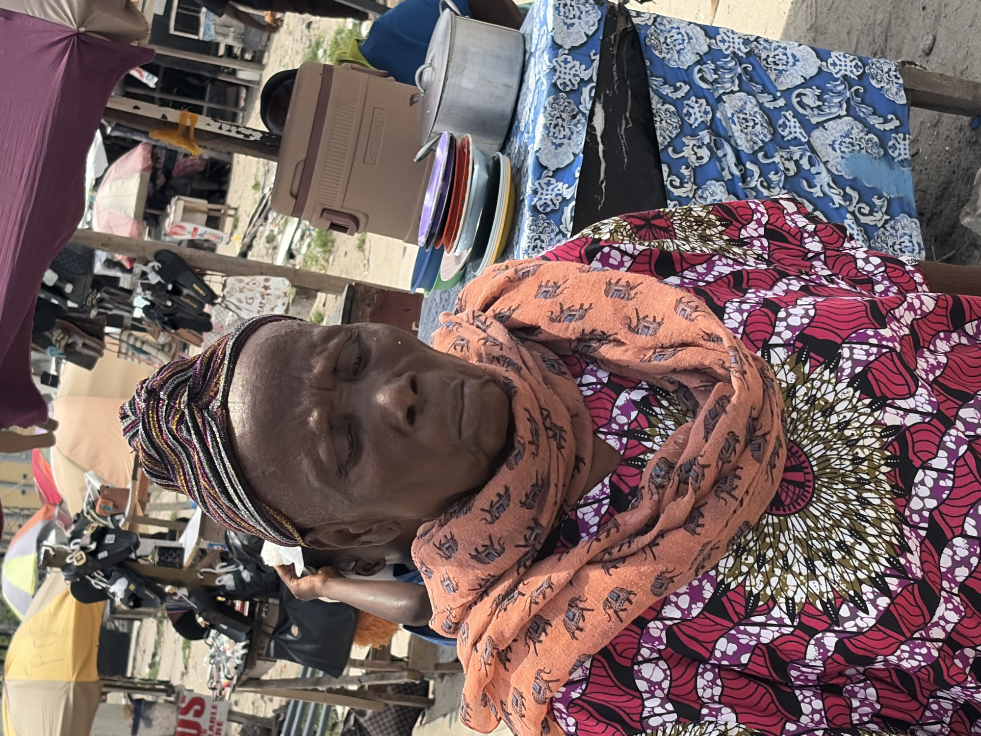 An elderly woman with brown skin and a multicolored headscarf is seated at an outdoor market stall in Igboefon, Nigeria. She wears a reddish-purple patterned dress and an orange scarf with elephant designs. The stall is covered with a blue patterned cloth and holds a metal pot, plates, and a cooler. Additional merchandise such as shoes and bags is displayed under umbrellas behind the woman. The ground appears sandy or dusty.