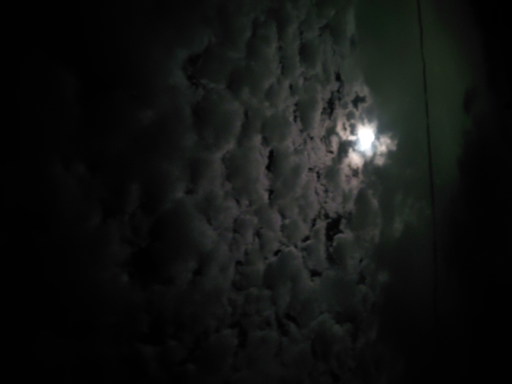 The image captures a dark, cloudy night sky, with a bright moon partially obscured by dense cloud formations. The perspective is upward-looking, showing a vast expanse of textured clouds filling most of the frame. The clouds are predominantly dark gray and black, with brighter white and light gray areas where the moonlight penetrates.
The moon is visible as a brilliant, glowing light source, its radiance diffused by the surrounding clouds, creating a soft, ethereal glow. This illumination highlights the contours and varying densities of the cloud cover, showcasing their lumpy, almost bubbly texture, characteristic of altocumulus or stratocumulus clouds.
In the upper right section of the image, a faint, dark vertical line can be discerned against a slightly greenish-tinged part of the sky, suggesting a possible power line or cable. No human activity or specific man-made objects are clearly identifiable, making the scene focused entirely on the natural elements of the night sky.
The time of day is unmistakably night, with the moon serving as the primary light source in an otherwise dark environment. The weather conditions are overcast and cloudy. While the location is contextualized as Santa Ana, Venezuela, there are no specific geographic landmarks or cues within the image itself to confirm this. No visible text is present. The image evokes a sense of natural mystery and quiet contemplation of the night sky.