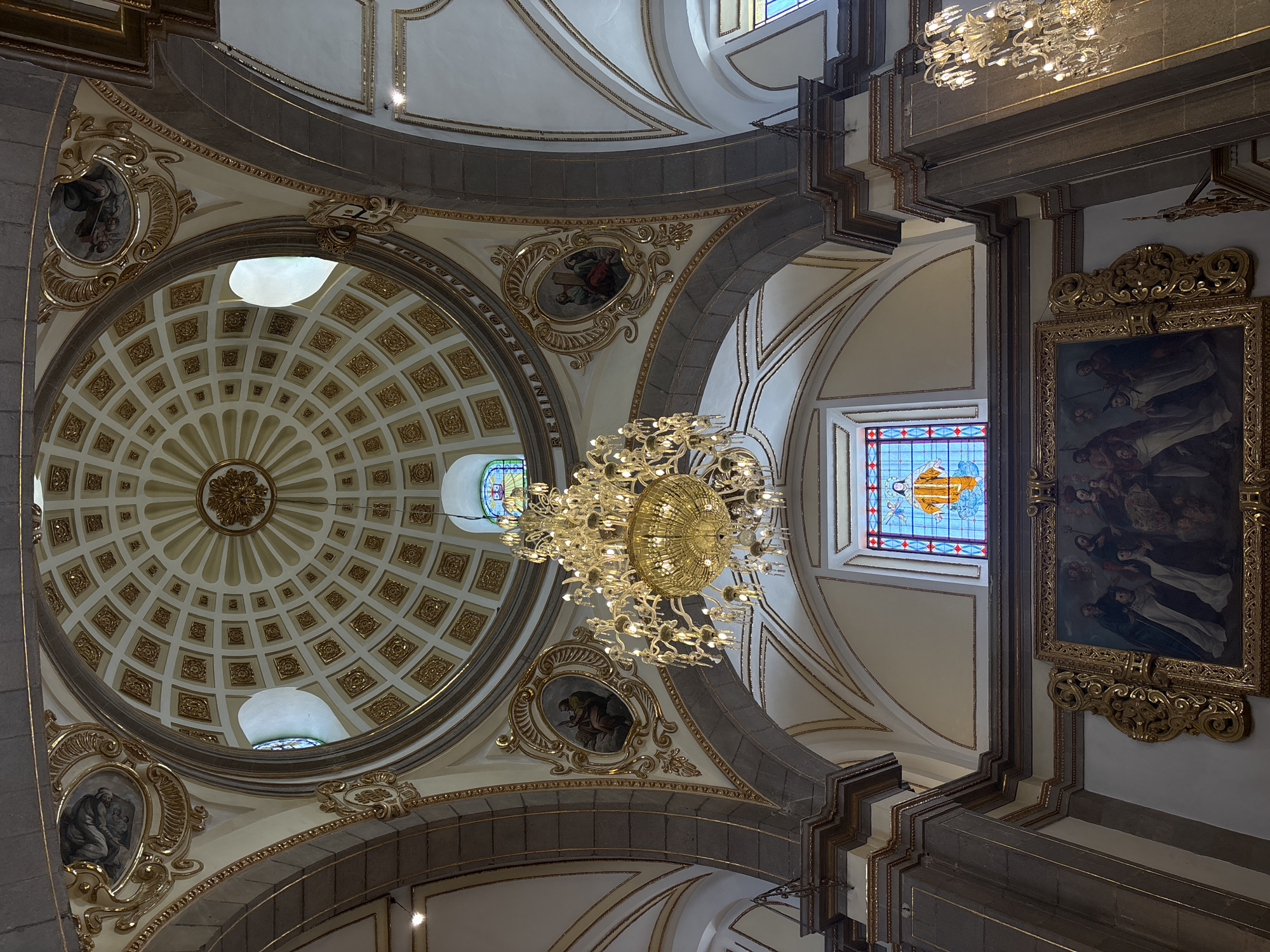 The image depicts the interior of a church in Puebla, Mexico. The perspective is looking upwards towards the ornate ceiling and dome. The dome is a circular structure with a pattern of concentric circles, each divided into smaller, square sections. These sections are decorated with gold accents. Light enters the dome through multiple small, round windows. A large, elaborate chandelier hangs in the center of the image, emitting a warm glow. The walls of the church are adorned with framed paintings and stained-glass windows. The architecture features arches and decorative molding. The color palette is dominated by whites, golds, grays, and muted colors from the stained glass.