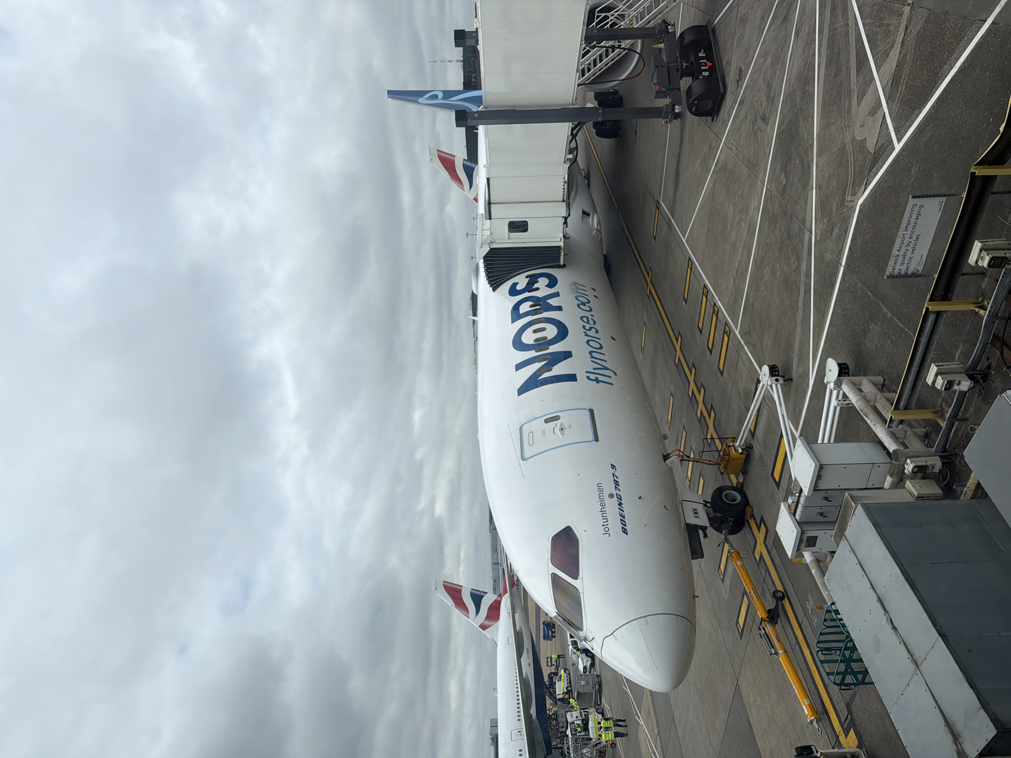 The image captures an aerial view of the nose of a Norse Atlantic Airways Boeing 787-9 aircraft, named "Jotunheimen," parked at an airport gate. The jet bridge is attached to the aircraft. The airline's logo, "NORS flynorse.com," is prominently displayed. In the background, a British Airways aircraft can be seen. The setting is Gatwick Airport in Crawley, United Kingdom, as indicated by the visible signage mentioning "Gatwick Airport improving air quality by encouraging electric vehicles." Ground crew members in yellow vests are visible near the aircraft. The weather appears overcast.