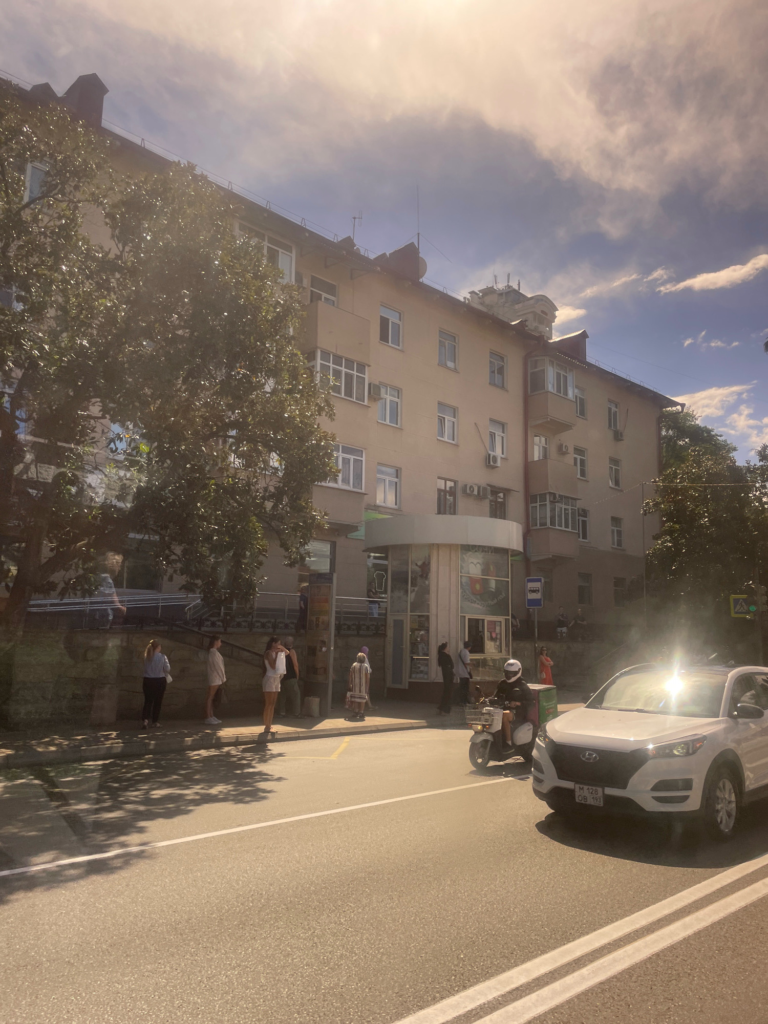 In Sochi, Russia, on a sunny day with a hazy sky and visible clouds, a street scene unfolds in front of a multi-story building. The building, painted in light beige and featuring numerous windows and balconies, houses a business on its ground floor with a curved entrance. People are gathered on the sidewalk in front of the building, some standing in line, while others are walking. A large tree with dense foliage casts shadows on the sidewalk and the street. A white SUV is parked on the right side of the road, its license plate visible. A person on a scooter with a green delivery box on the back is driving past the SUV. The street is paved and marked with white lines. The overall impression is of a typical urban street scene in Sochi, with everyday activities taking place under a bright sky.