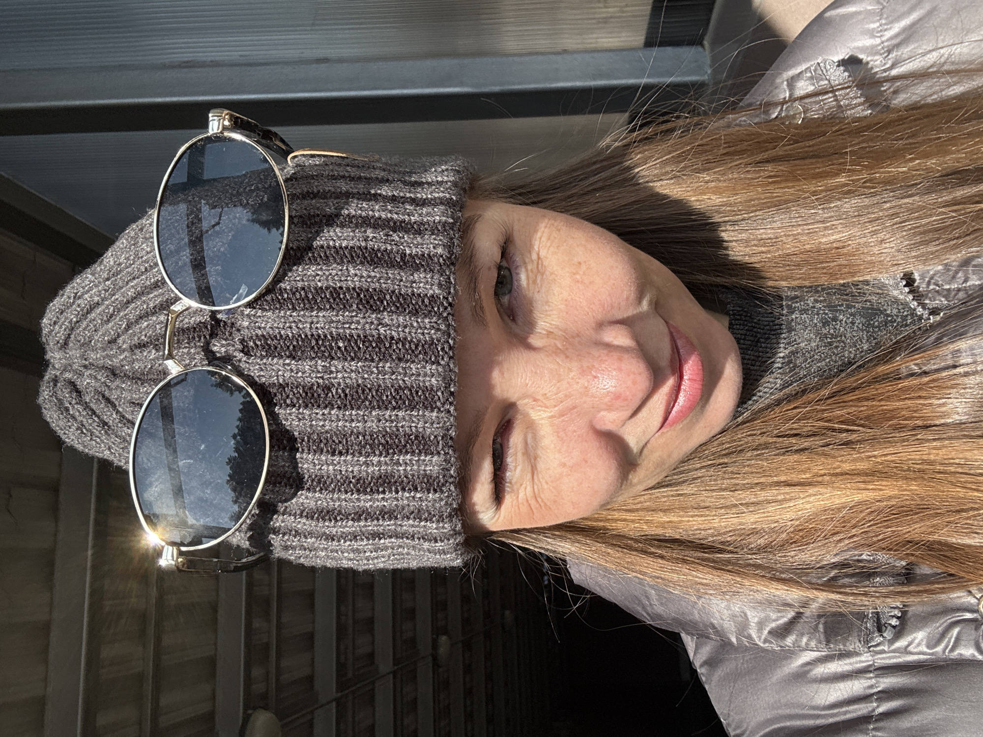 The image depicts a close-up portrait of an individual, identified as being in Прикамский, Russia. The subject is an adult female with long, light brown hair, wearing a dark grey ribbed knit beanie with oval-framed sunglasses perched on top. The sunglasses have silver frames and dark lenses, reflecting trees and a bright sun flare on the left lens. She is wearing a shiny, grey quilted jacket over a dark grey knit sweater. Her left eye is winking, and she exhibits a slight smile with closed lips. Freckles are visible on her nose and cheeks. Strong direct sunlight illuminates her right side, hair, and the sunglasses, creating shadows on the left side of her face. The background consists of a dark, tunnel-like structure with visible beams and a light fixture to the upper left, transitioning to a sunlit, vertically paneled wall or structure on the right.
