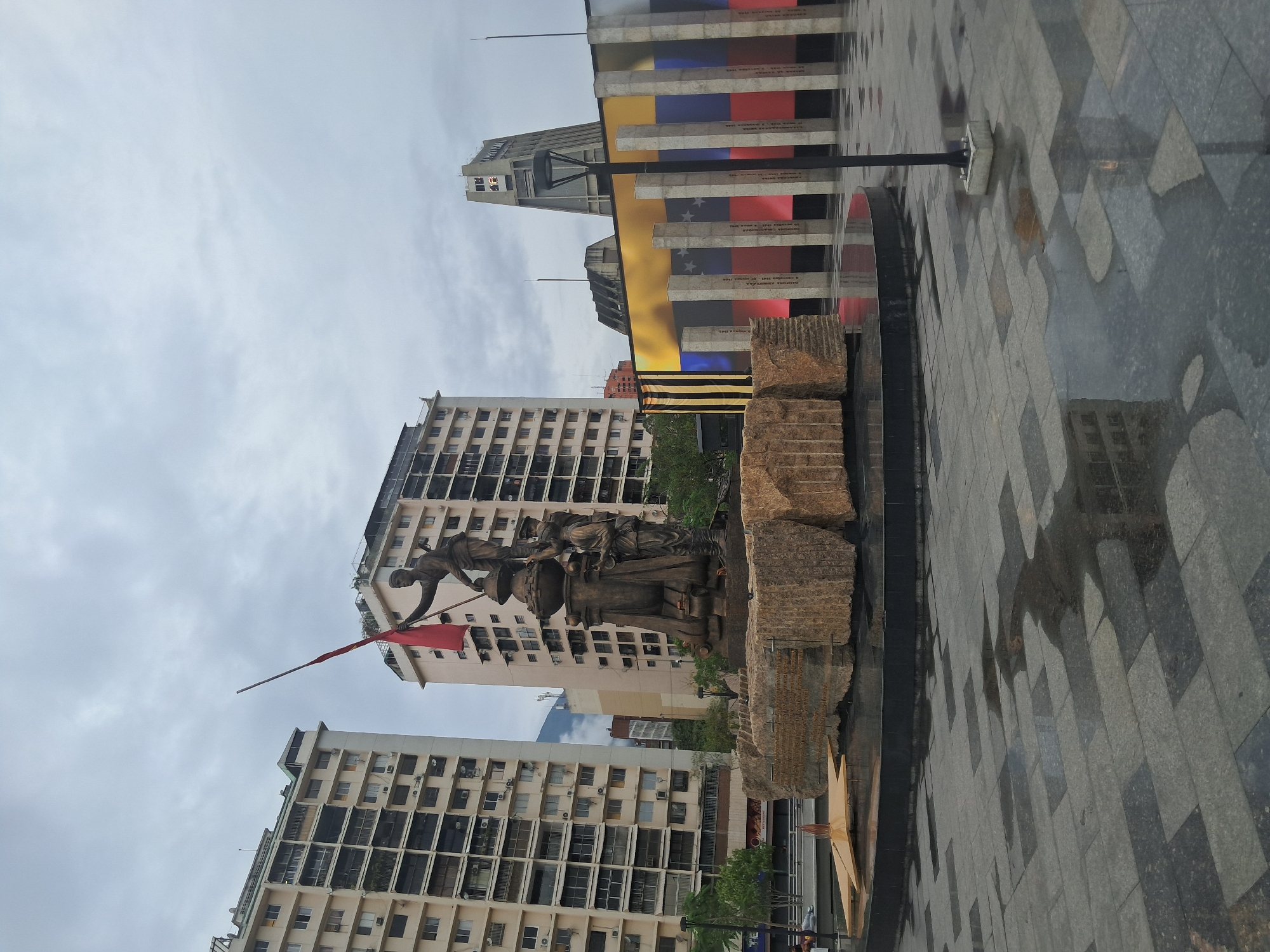 The image shows the Monument to the Heroes in Caracas, Venezuela, on a cloudy day. The monument consists of a bronze sculpture of figures carrying a flag, set on a stone base. In the background are apartment buildings and a tower with an image of the Venezuelan flag. The surrounding pavement is made of gray and black tiles, with reflections suggesting it may be wet.