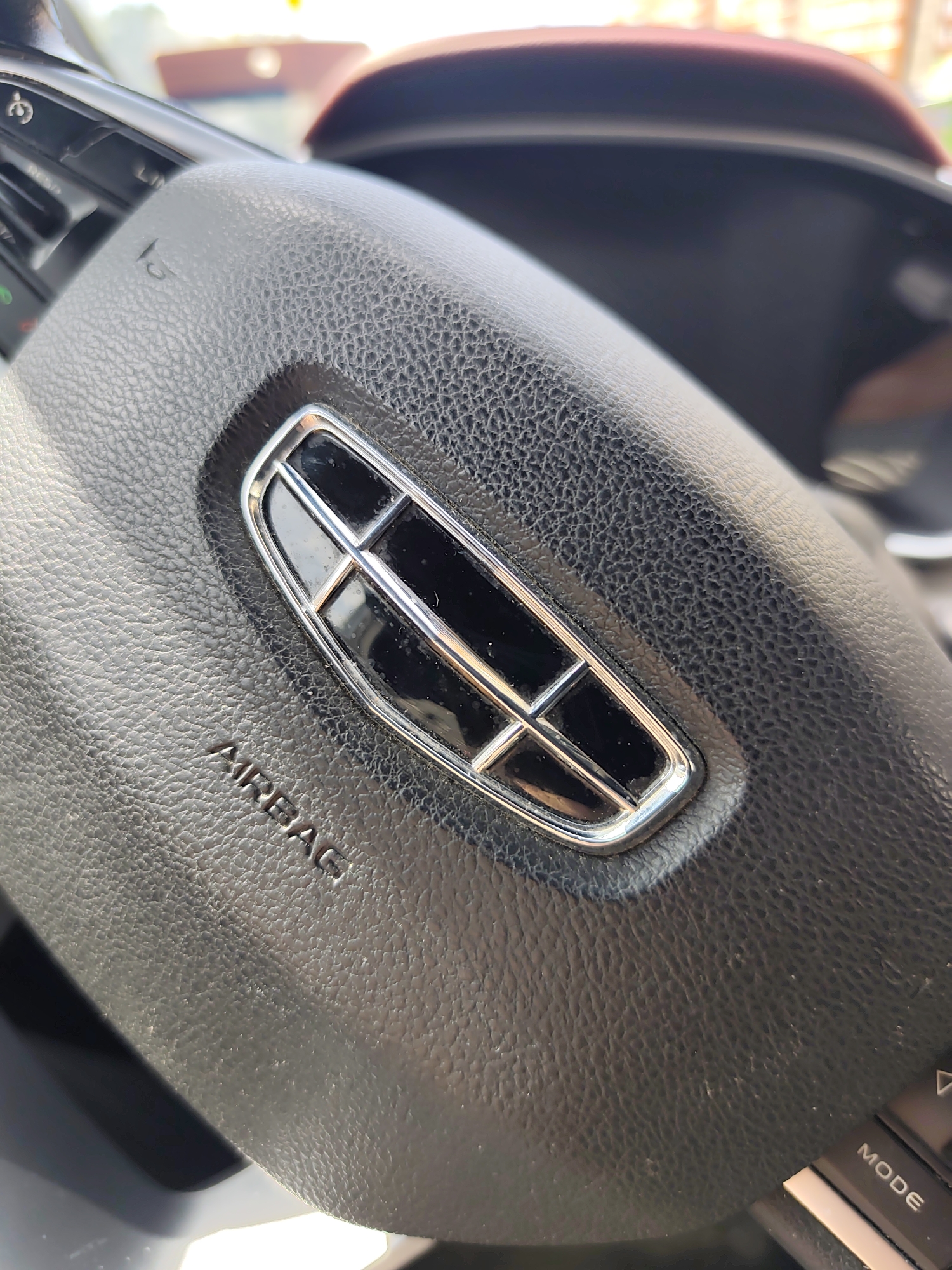 The image shows a close-up view of a car's steering wheel. The steering wheel is black and textured, with a metallic emblem in the center. The emblem has a dark, rectangular shape divided by thin chrome lines. Below the emblem, the word "AIRBAG" is embossed into the leather. To the left of the emblem, on the steering wheel's controls, the word "LIMIT" is visible, along with a horn symbol and other buttons which are partially obscured. To the right of the emblem, the word "MODE" is visible on another set of buttons. The background is out of focus, but appears to be the interior of a car, with a glimpse of a dashboard and possibly the windshield. The lighting suggests it is daytime, with sunlight creating some reflections on the steering wheel. The scene is likely set in Ryazan, Russia, as indicated by the location context. No people are visible in the image, and the focus is entirely on the steering wheel and its details.