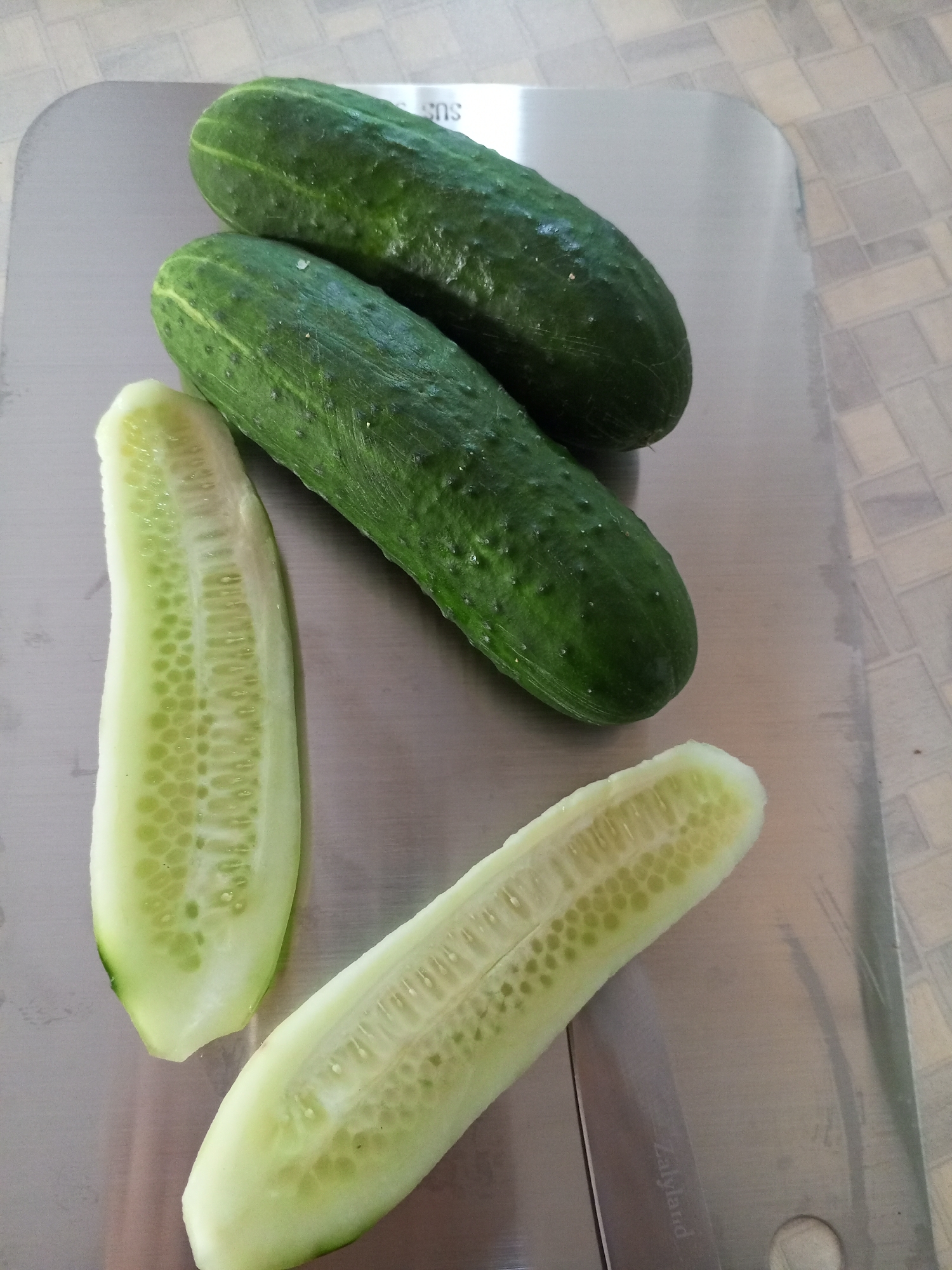 Three whole cucumbers and two halves of a cucumber are arranged on a stainless steel surface. The surface has a brushed metal finish and appears to be a cutting board or tray. Two whole cucumbers are positioned at the top of the frame, with one slightly overlapping the other. A third whole cucumber lies below them, running diagonally across the surface. Two cucumber halves, showing the internal seeds and flesh, are placed towards the bottom of the frame. The background consists of a tiled surface with a checkerboard pattern in muted tones. There are no people or animals visible in the image. The lighting is neutral, with no strong shadows. The image's origin in Vilyuysk, Russia, is a detail not visually represented in the provided image content.
