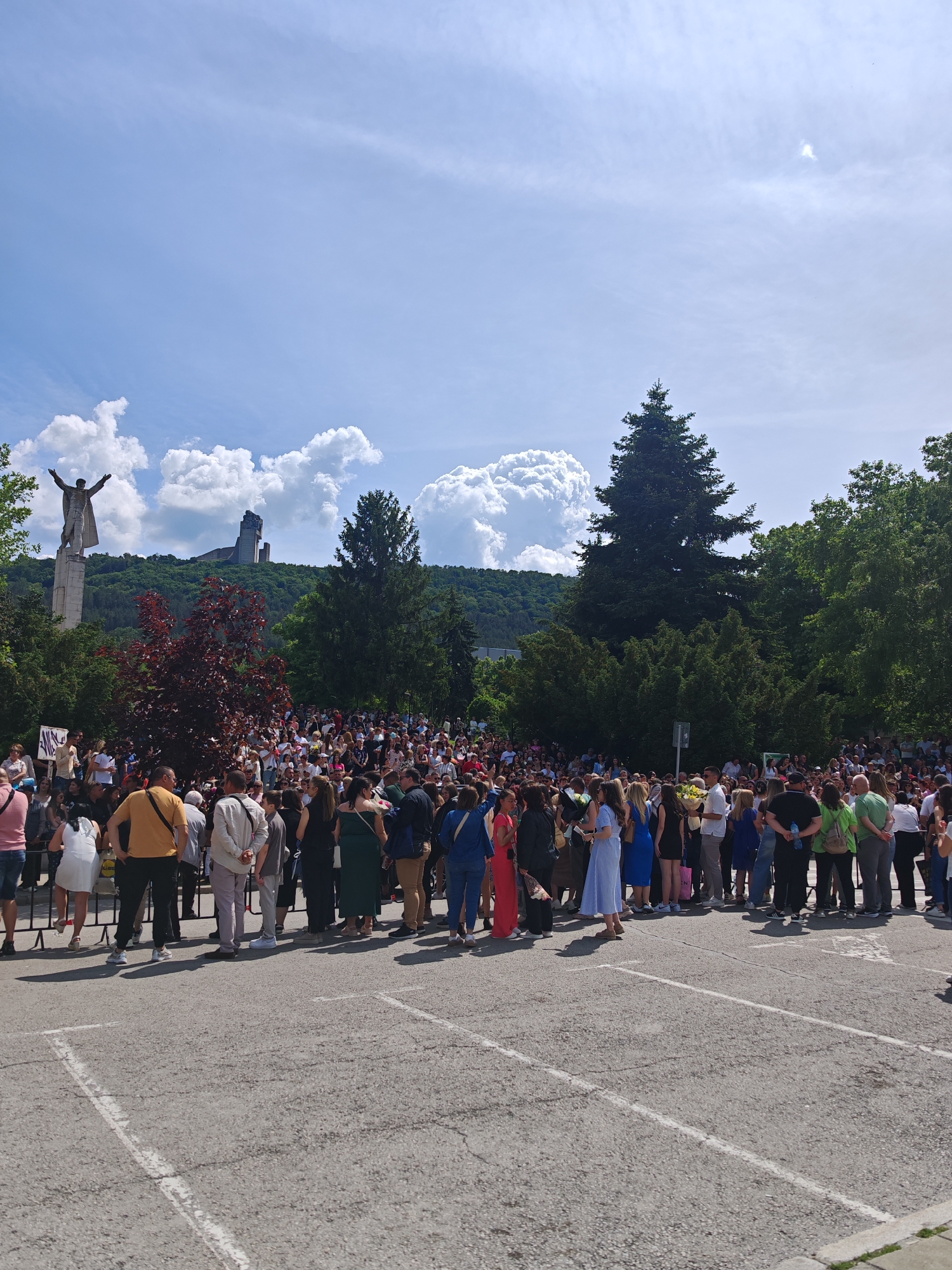 A large crowd of people stands on a paved parking lot, behind a low metal barrier, facing away from the viewer. The people are dressed in a variety of casual and formal attire. Many appear to be holding flowers. In the background, there is a statue of a man with arms outstretched, and behind that, a monument sits atop a hill. Green trees and bushes fill the landscape. Above, the sky is blue with puffy white clouds. This is likely a graduation ceremony. The location is Shumen, Bulgaria, with the Founders of the Bulgarian State monument visible in the background.