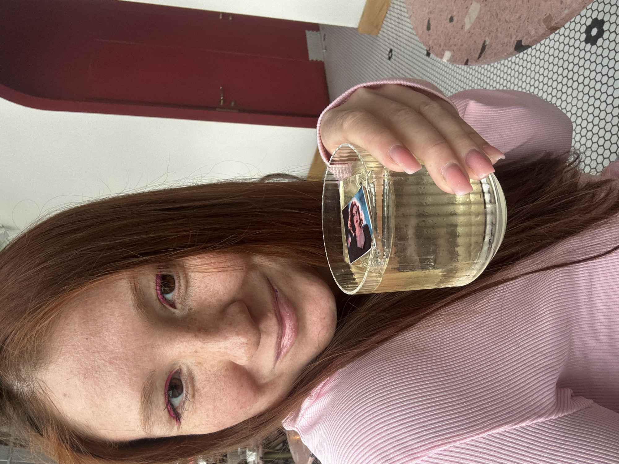 A young woman with red hair and freckles, wearing pink eyeliner and a pink ribbed shirt, is holding a glass of clear liquid in her right hand. A small photograph is affixed to the inside of the glass. The background includes a red door, white wall, and a tiled floor with a pink pattern. The location is likely an indoor setting. The specific location in Nizhny Novgorod, Russia is not discernible from the image.