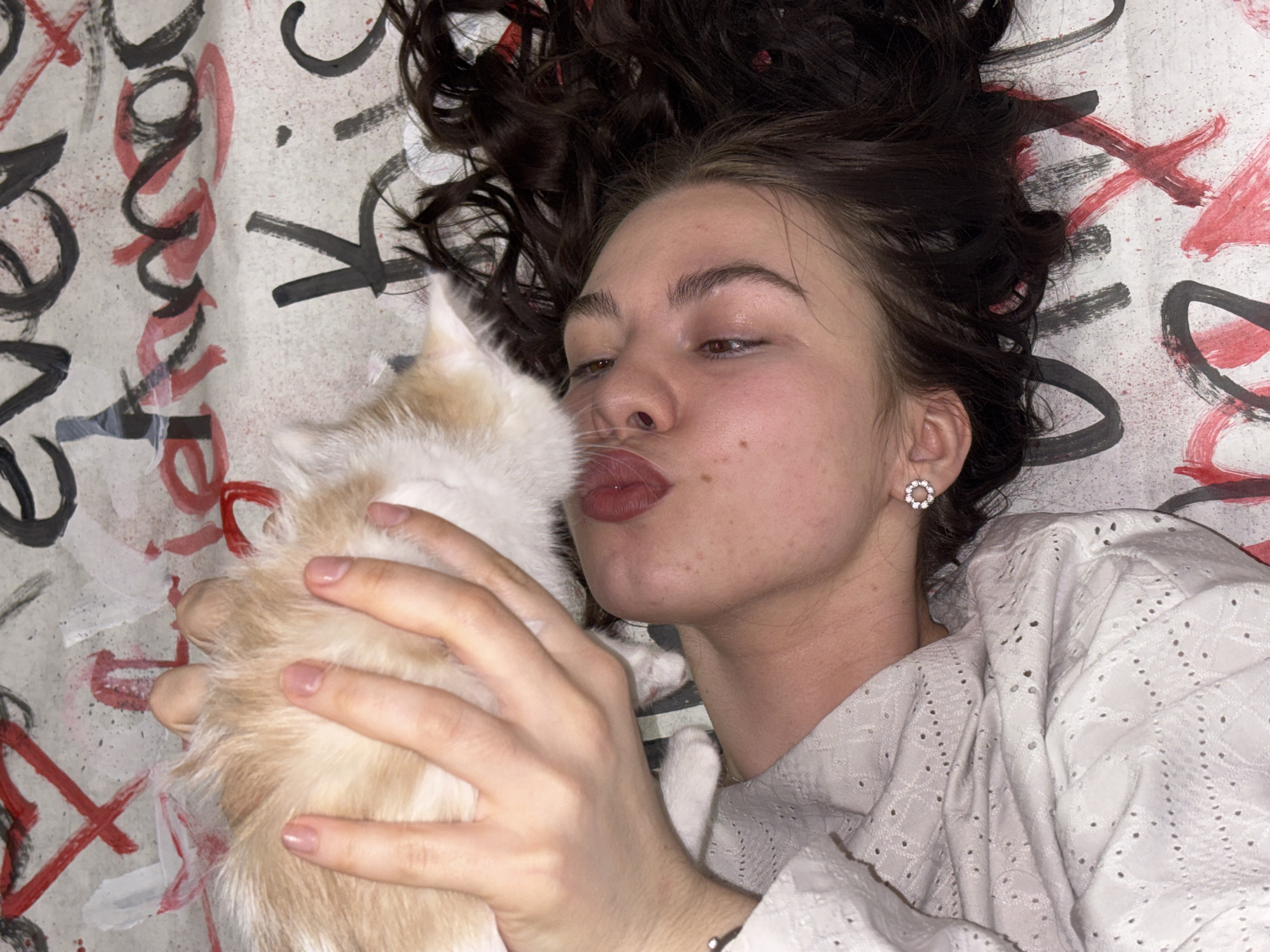 A young woman, likely in her late teens or early twenties, is depicted lying on her back, looking upwards. Her dark, curly hair is spread out around her head on a patterned surface. She is holding a small, predominantly white and light orange/ginger cat close to her face, with her lips puckered as if preparing to kiss it. The cat's head is turned towards her.
The woman has fair skin with visible freckles and natural-looking eyebrows. She is wearing a light-colored top, possibly white or cream, which features an eyelet or embroidered texture. A sparkling, circular stud earring is visible in her right ear.
The background is a textile surface, possibly a bedspread or blanket, decorated with a bold, graffiti-style pattern. This pattern includes black and red brushstrokes forming abstract shapes and stylized letters. Fragments of text, such as what appears to be "DORNE" or "DONNA" in black and red, are partially visible and upside down. Multiple red "X" shapes are also prominent.
The image captures an intimate and affectionate moment between the woman and her pet, set indoors with soft, even lighting. The location context provided is Perm, Russia, but this is not visually evident from the image itself.