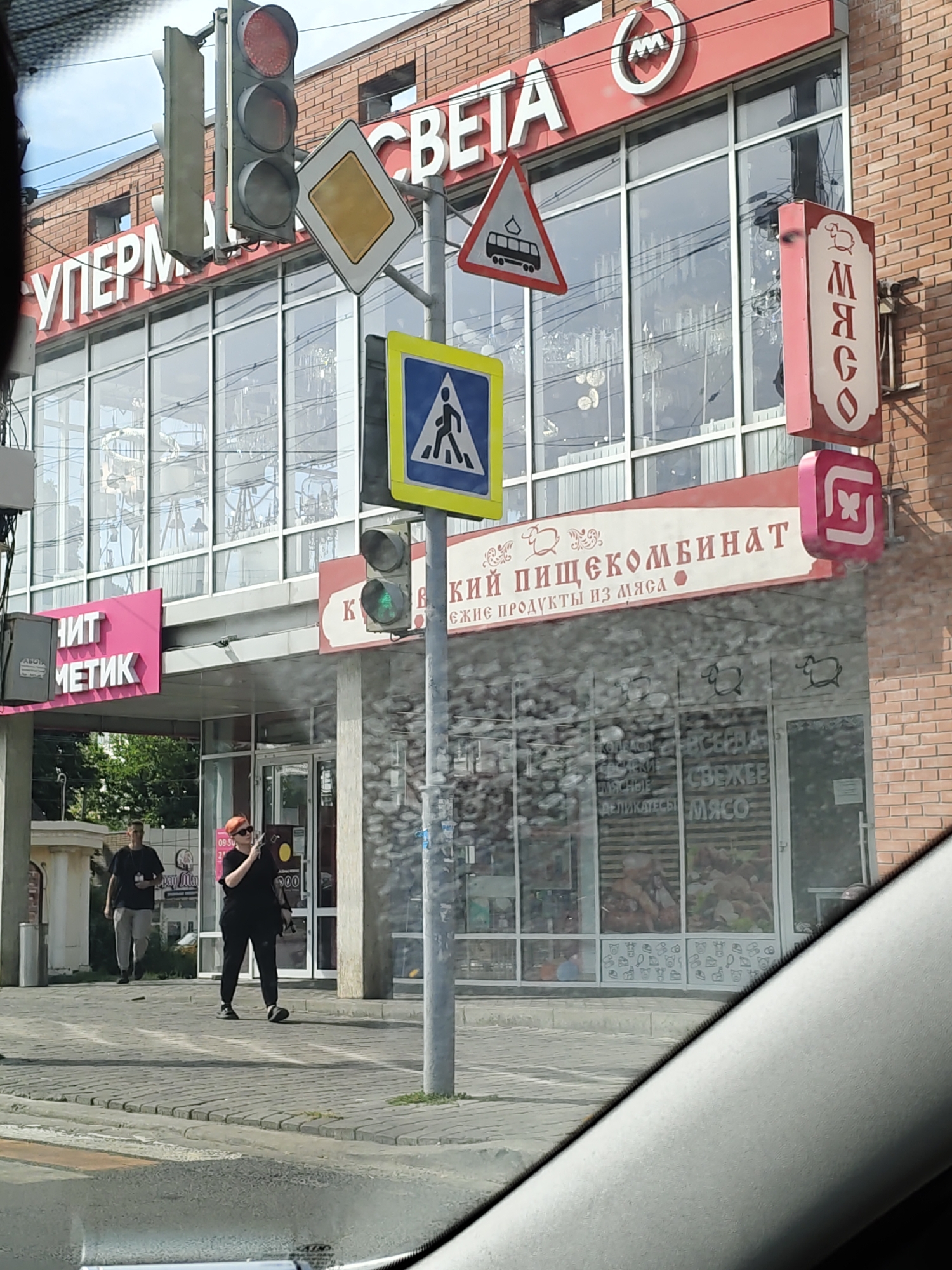 The image depicts a street scene in front of a brick building in Novocherkassk, Russia, featuring a supermarket and a meat products store. A traffic light shows a green signal, and various road signs, including a pedestrian crossing sign and a diamond-shaped priority sign, are visible. The building's facade displays multiple signs with Russian text, indicating a supermarket and a "Meat Products Combine." Inside the store, through the glass windows, shelves stocked with meat products are discernible. Two individuals are present on the sidewalk; one man is walking away from the camera, while a woman, holding a phone, stands facing the building. The environment suggests an urban setting with paved sidewalks and a road.