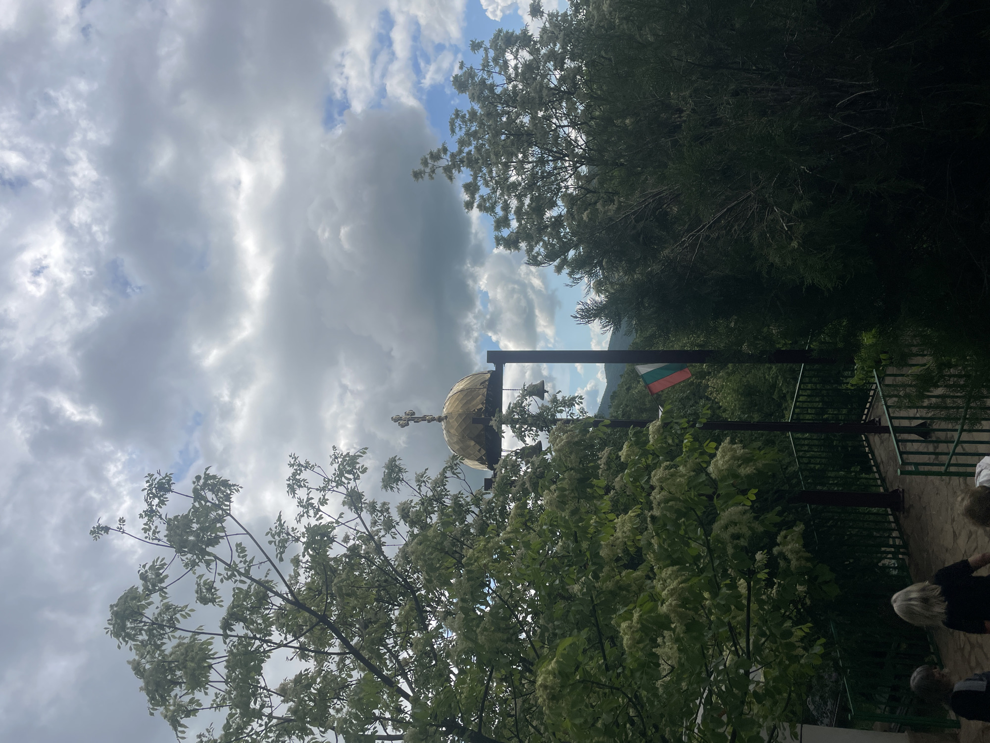 A cloudy day, partially obscured by dense foliage, reveals a glimpse of the Novo Selo, Bulgaria, landmark. A golden dome, capped with a cross, rises above the trees, suggesting the presence of a church or religious site. Adjacent to the dome, a structure bears the Bulgarian national flag. A paved walkway and a fence appear, indicating a viewing point or gathering area, where several people are present.
