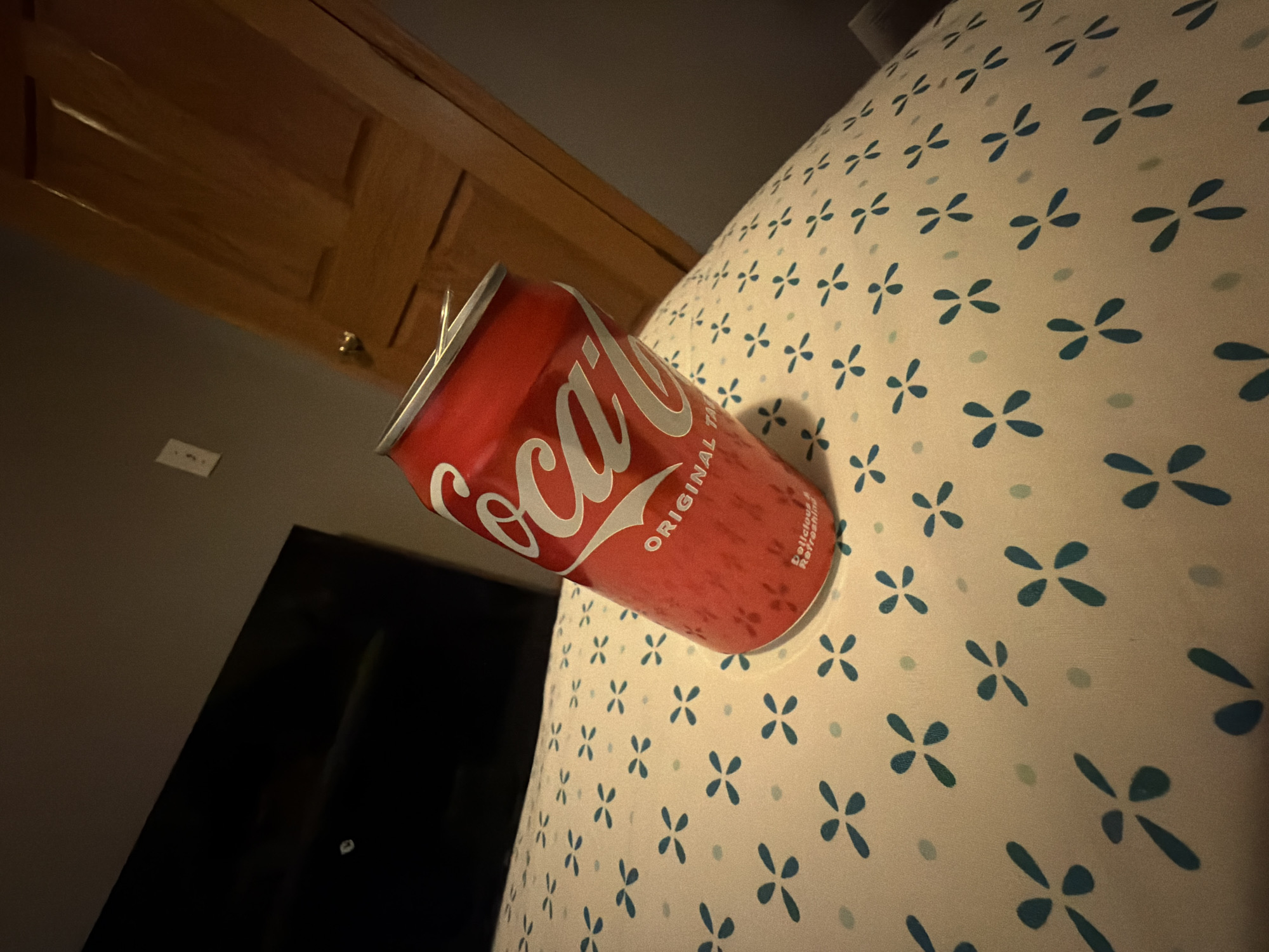 A red Coca-Cola can sits on a white patterned surface, likely a bedspread, which is decorated with small blue floral motifs and light green dots. The can is slightly tilted, showing condensation on its surface. In the background, a dark, flat screen television is visible on the left, and a wooden door with a doorknob is on the right. The scene is dimly lit, suggesting an indoor setting. The location is the city of Ham Lake, United States of America.