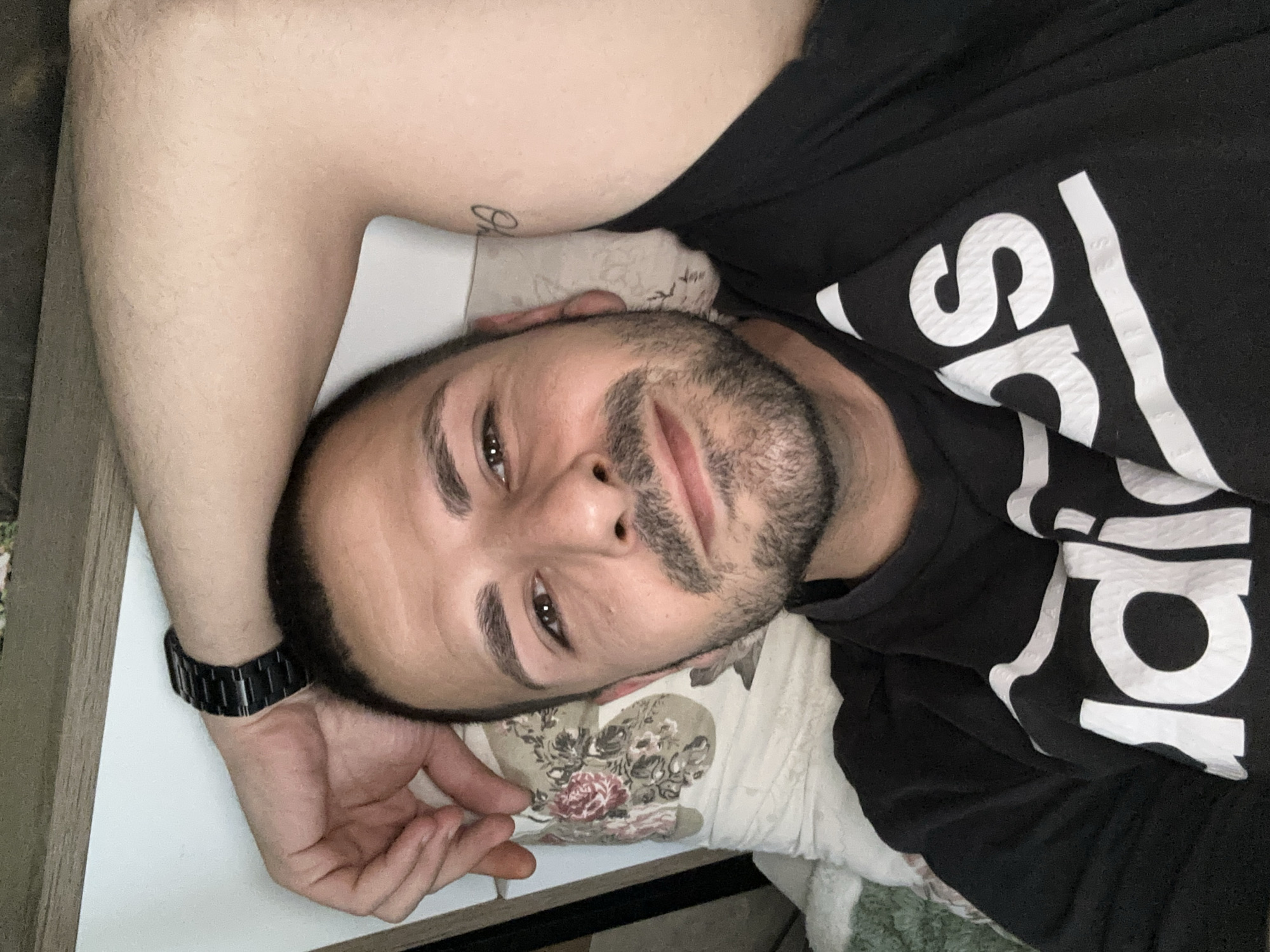 A selfie shows a man with a dark beard and short dark hair, wearing a black Adidas t-shirt and a black metal watch on his left wrist. He is lying down, with his head on a floral-patterned pillow and his arm raised above his head, revealing a tattoo on his upper arm. He is looking at the camera. The background is blurred. There is no indication of the location being Slavyani, Bulgaria.