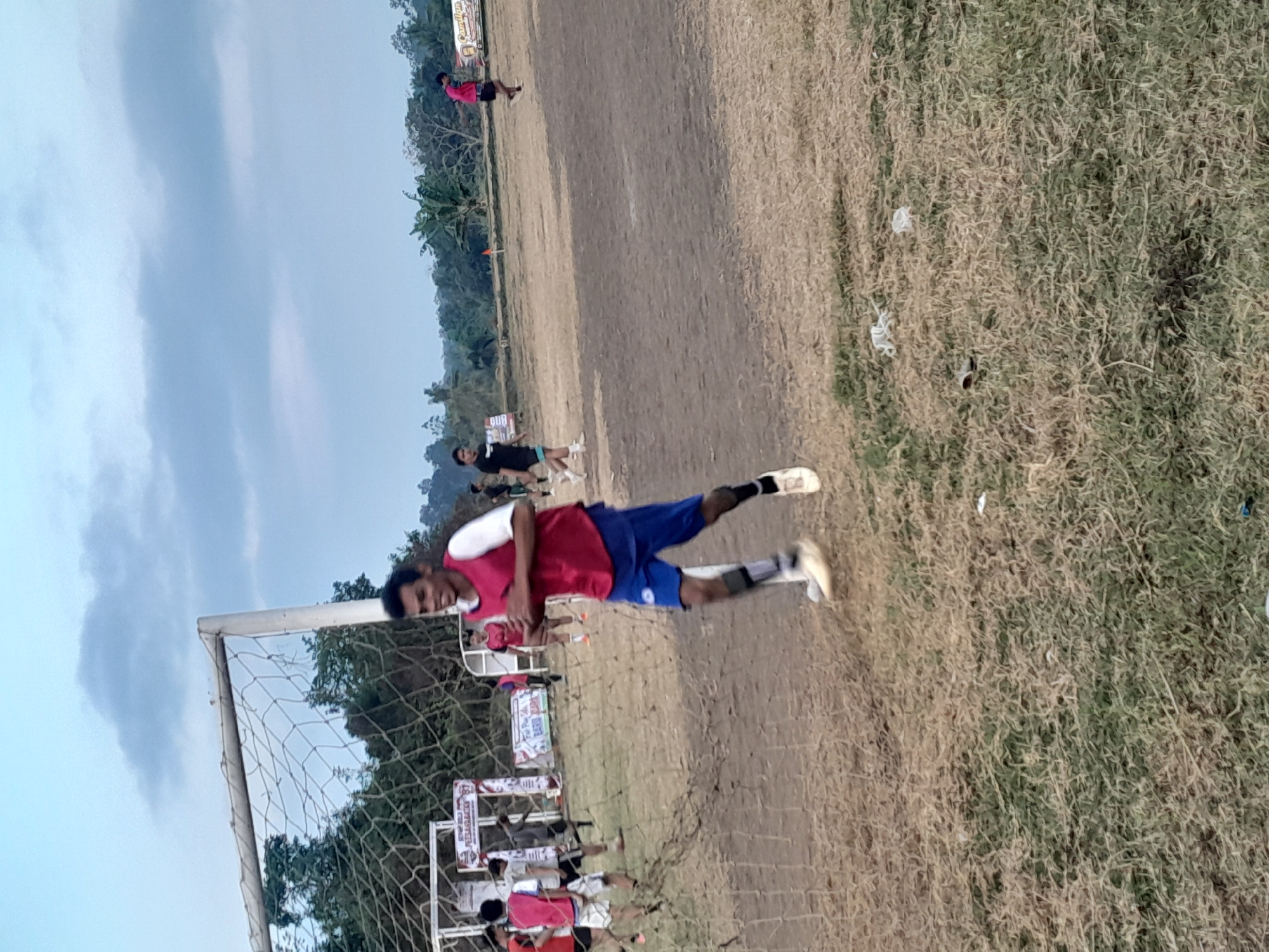 A group of young men are playing soccer on a dirt field, with a goal and net visible in the foreground. The men are wearing athletic clothing, with some in red and white jerseys and others in black. The field is dry and dusty, with sparse grass. In the background, there are trees and a cloudy sky. The scene appears to be in a rural or suburban area. Based on visual cues and the information provided, this scene is likely located in Genteng, Indonesia, as indicated by the banners with Indonesian text visible on the field.