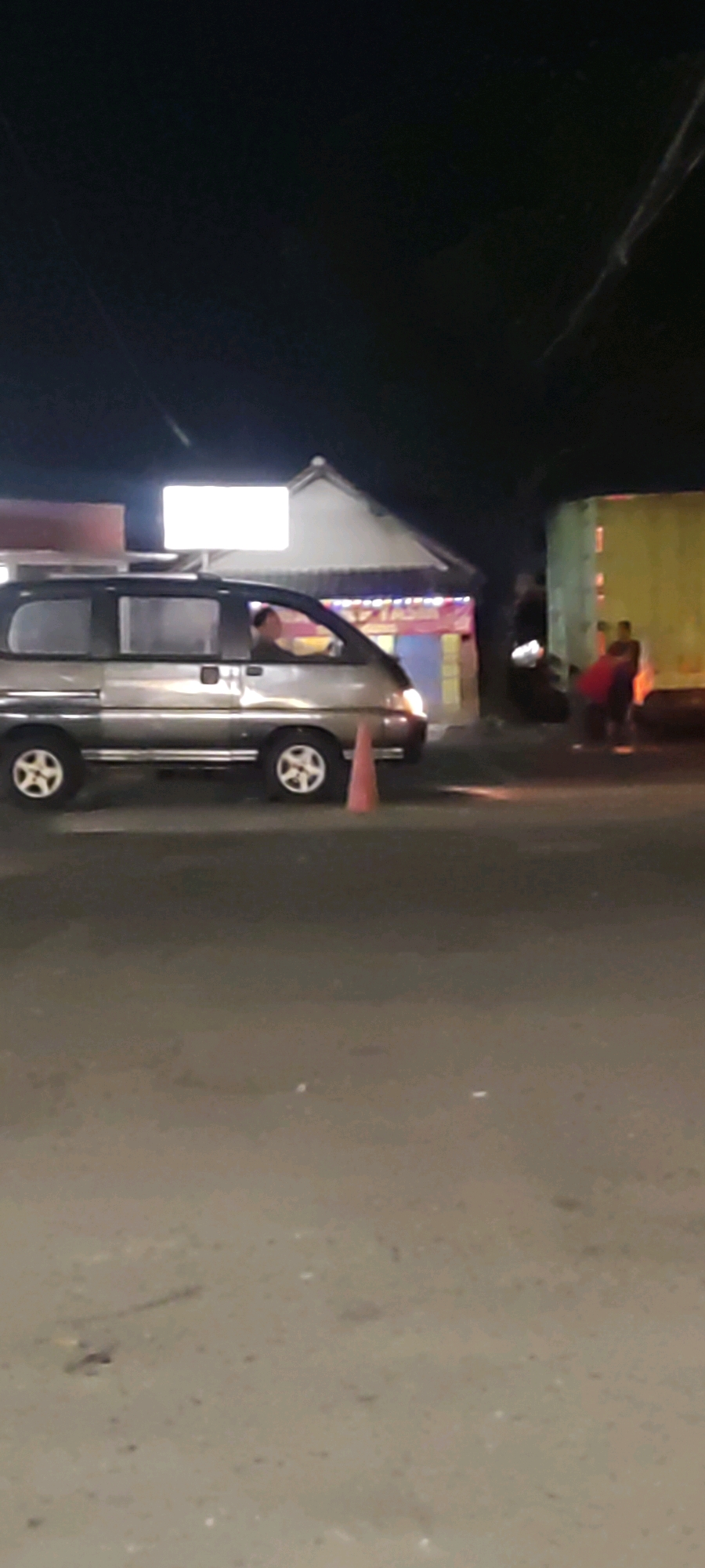 In the city of Pelayangan, Indonesia, at night, a silver van is parked on a road. The van is a passenger van with tinted windows. A person is visible inside the van, in the driver's seat. In front of the van, a traffic cone is placed on the road. In the background, there is a building with lights and some people standing near a truck. The environment appears to be a street with some commercial establishments.