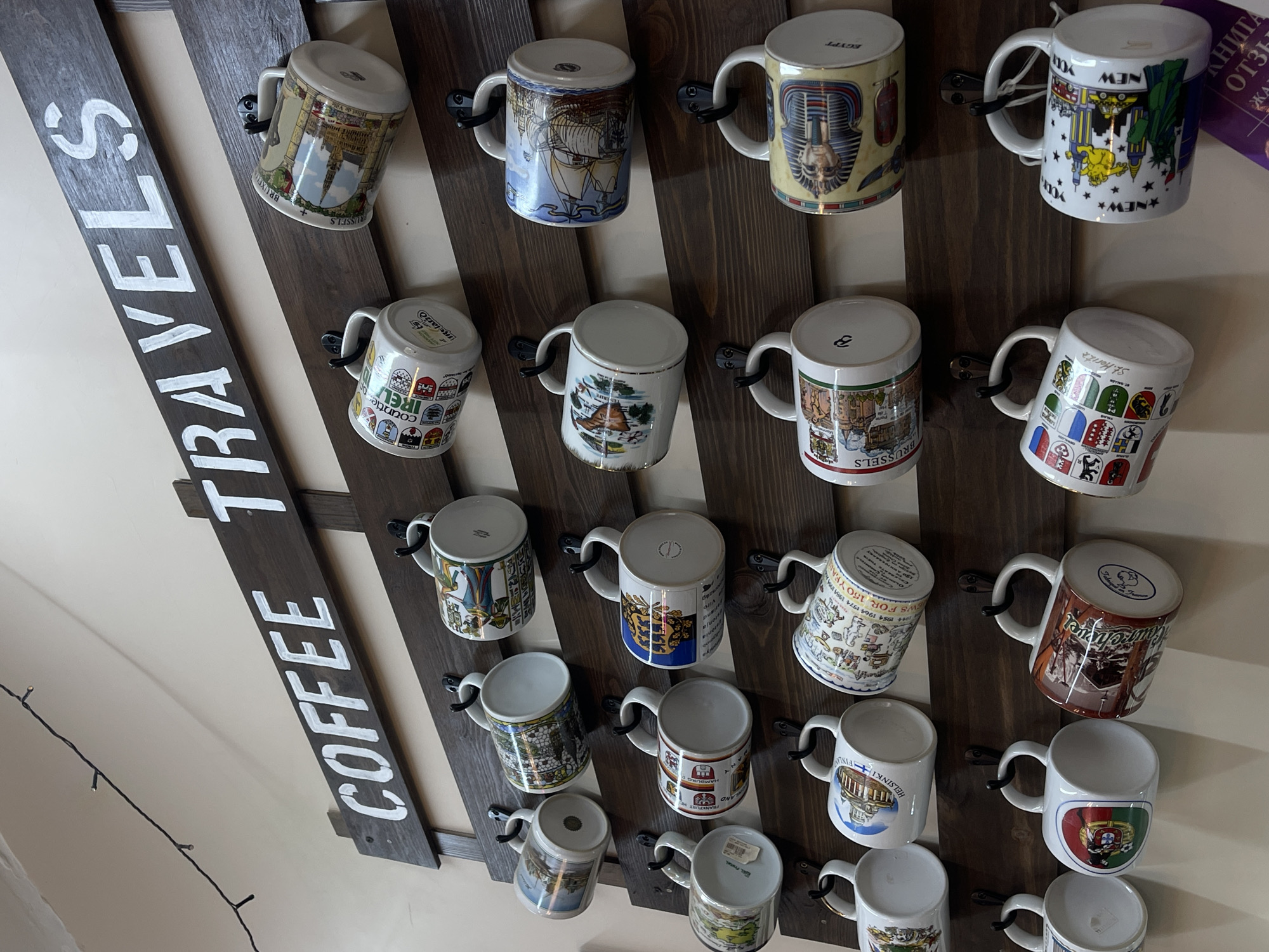 The image shows a wall-mounted display of coffee mugs in what appears to be a souvenir shop or cafe in Ostrov, Russia. The display is constructed from dark wood planks arranged horizontally, with black metal hooks attached to hold the mugs. A dark wooden plank at the top of the display has the words "COFFEE TRAVELS" painted in white stencil letters.
There are multiple rows of mugs, each adorned with various designs, likely depicting places and landmarks from around the world, characteristic of travel souvenirs. The mugs are primarily white ceramic with colorful illustrations. In the bottom right corner, a purple tag with Russian text and "ЗАП" is visible, suggesting it is a tag for a visitor's book or souvenir item. The background is a light-colored wall with a string of small, dark fairy lights running along the top left. No people are visible in the image.