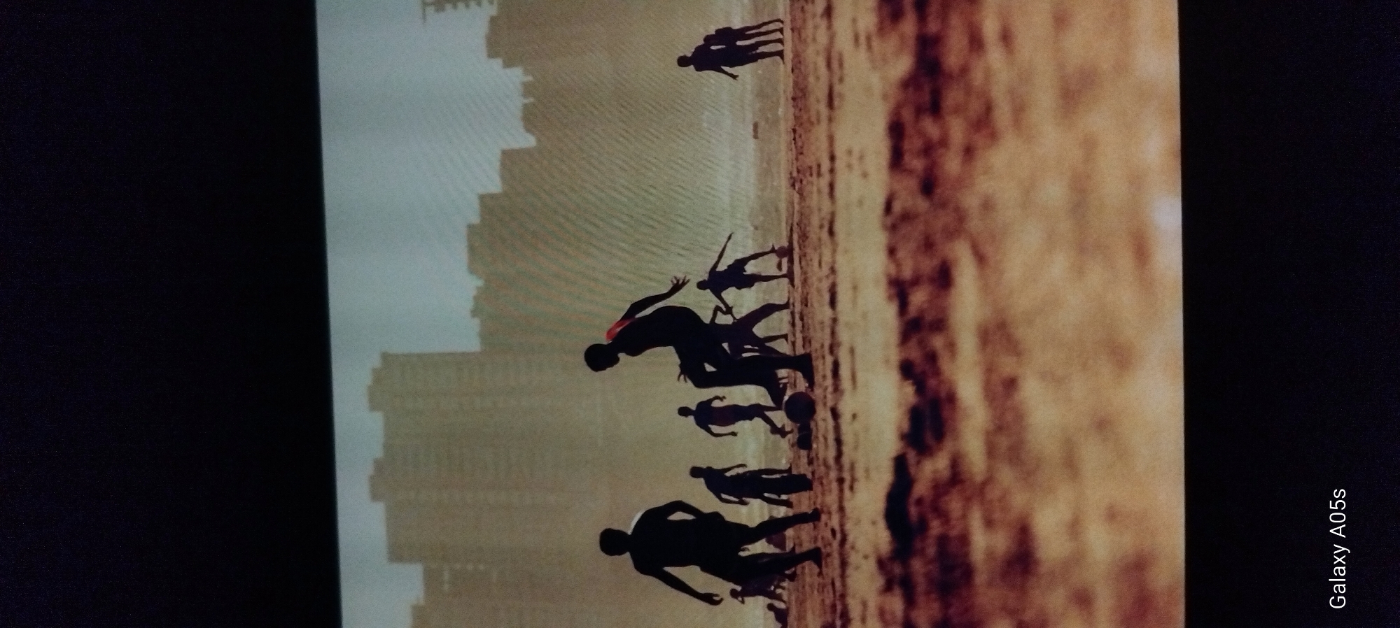 The image displays a vibrant scene of several individuals, likely children or young adults, playing football (soccer) on what appears to be a sandy or earthy ground, possibly a beach or a dusty field. All figures are rendered as dark silhouettes against a bright, hazy sky, suggesting either sunrise or sunset. Their dynamic poses – running, kicking, and jumping – convey energetic movement and engagement in the game. A football is visible near the feet of some players.
In the mid-ground, a body of water is visible, merging into the distant horizon with the hazy sky. Further in the background, a distinctive silhouette of tall buildings or a city skyline can be seen against the soft, diffused light of the sky. The light source is behind the subjects, creating stark, dramatic outlines of the figures. One player has a faint hint of red visible on their clothing. The overall mood is one of active recreation and youthful exuberance.
On the far right edge of the original image (which appears to be a photograph of a screen), the text "Galaxy A05s" is visible in white, indicating the device that captured the image being displayed. While the provided context is Zavoy, Bulgaria, the image itself shows a scene that could be typical of many coastal or lakeside areas with a distant urban setting, without specific identifying landmarks for that location.