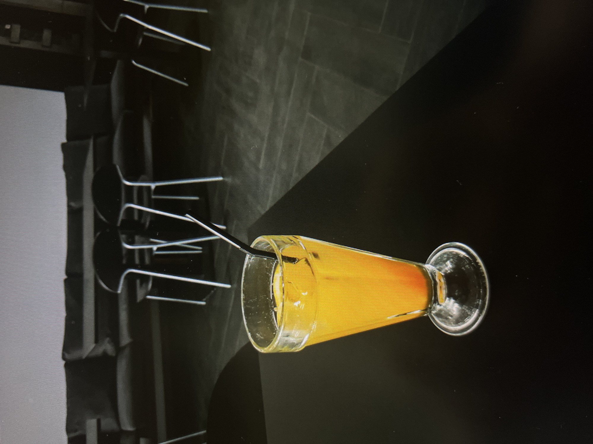 A tilted, high-angle shot depicts a glass of orange juice with a black straw, placed on a dark table alongside stacked chairs in a location that resembles a restaurant interior. The image is in monochrome except for the juice. The table is diagonally divided, one side being darker than the other. The chairs are visible in the background with their legs extending upward. Minsk, Belarus is not directly discernible from the image.