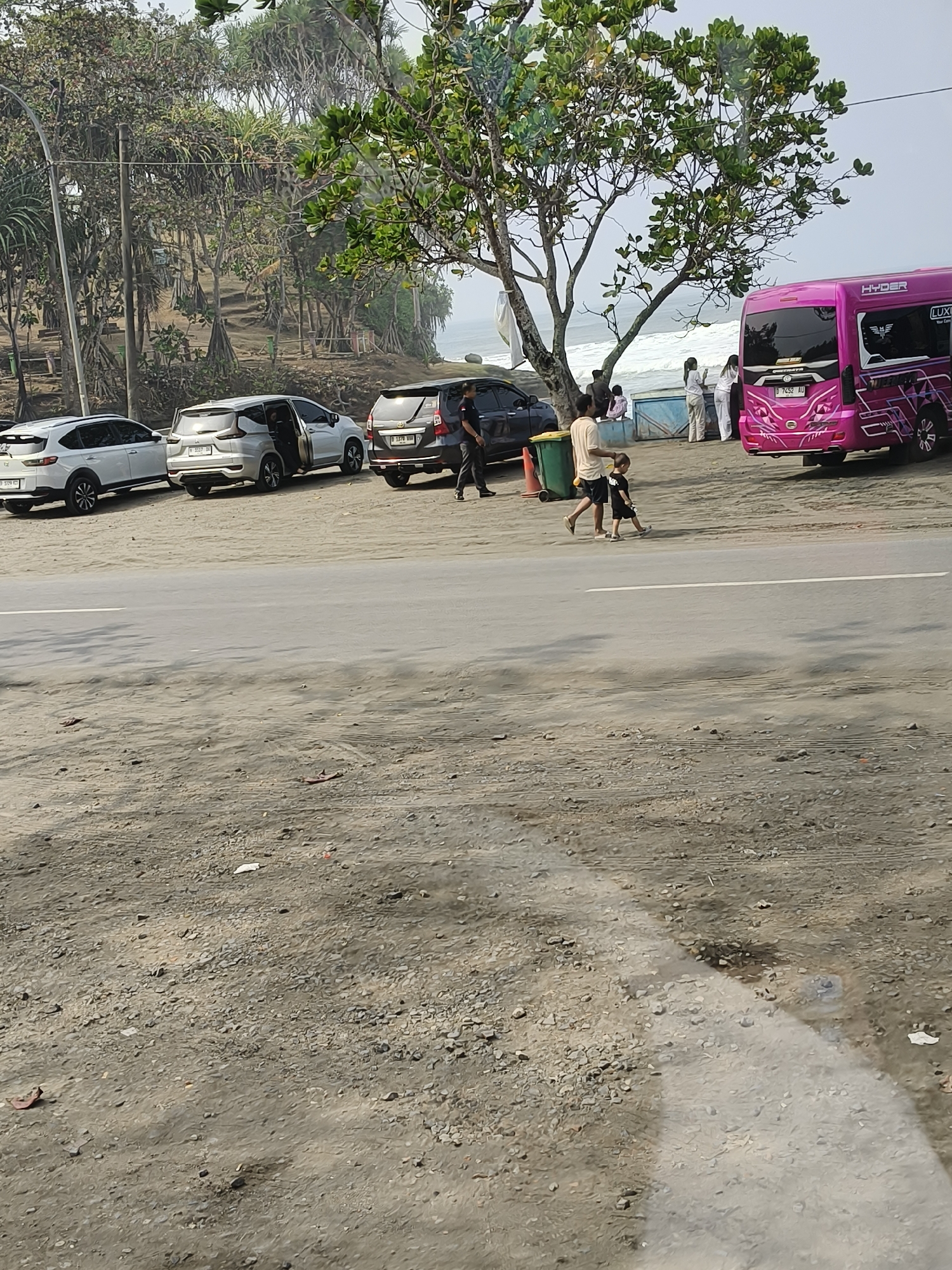 The image depicts a roadside scene at Desa Ciliang, Indonesia, with a body of water, possibly the ocean, visible in the background. Several vehicles are parked along the side of a road, including a white SUV, a silver minivan with its door open, and a dark-colored SUV. A vibrant pink minibus with "HYDER" and "LUXY" branding is also present. In the foreground, two people, an adult and a child, are walking away from the camera. The adult is wearing a light-colored shirt and shorts, while the child is in a dark t-shirt and shorts. The environment appears to be a coastal area, with sandy ground and vegetation, including trees, in the background. The sky is overcast.