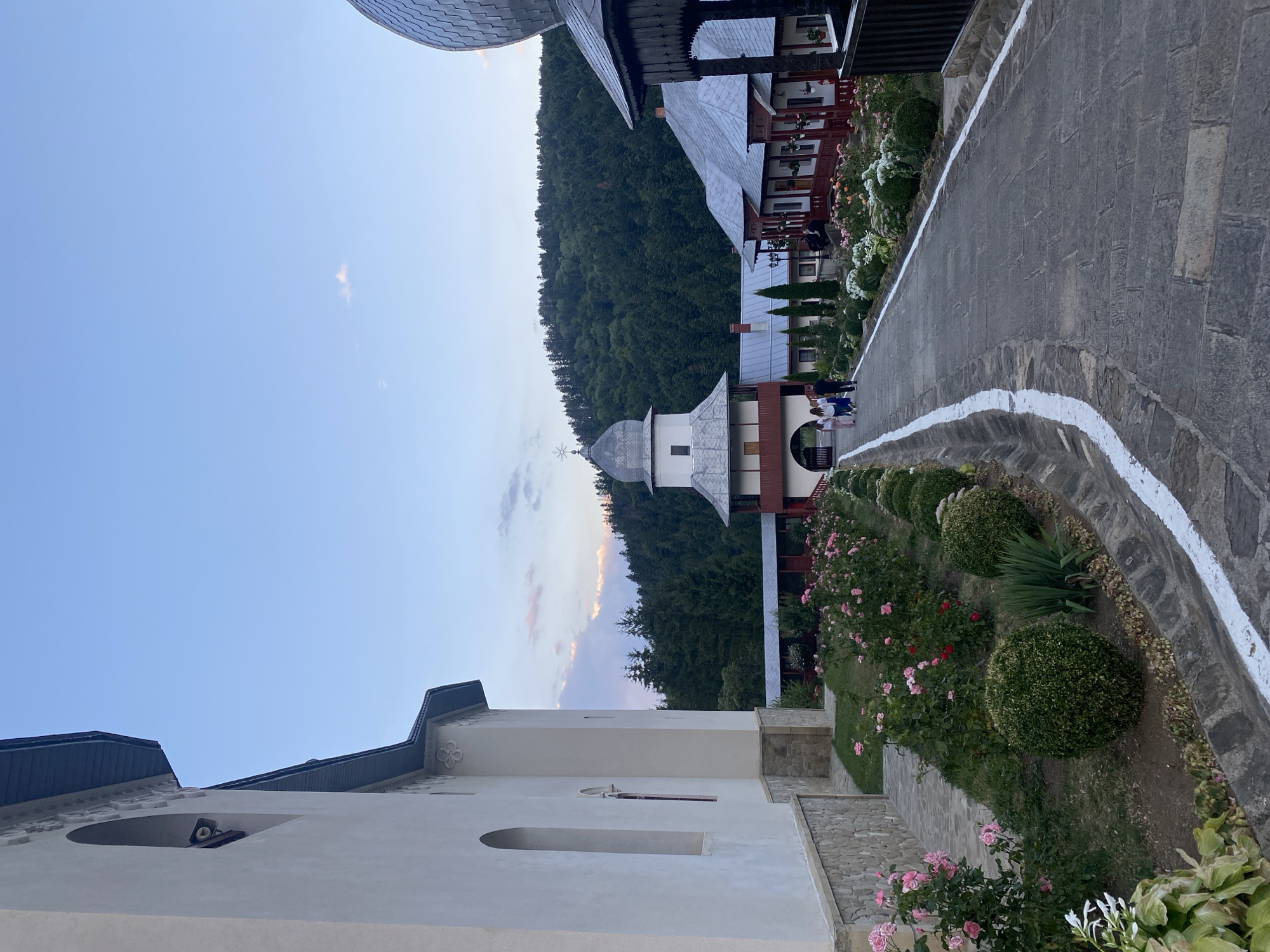The image depicts a pathway leading through a monastery complex in Crăcăoani, Romania. On the left, a section of a white building with arched windows and stone detailing is visible. The pathway is paved with grey stones and bordered by a drainage ditch painted with white markings. To the right of the pathway, a garden features rows of blooming pink roses and meticulously shaped green bushes. In the background, several traditional Romanian monastery buildings with red accents and grey roofs are arranged around a central courtyard. A prominent bell tower with a silver dome stands to the left of the buildings. A dense, green forest covers a hillside in the far background under a pale blue sky with wispy clouds. A few people are walking along the pathway, appearing to be visitors or residents of the monastery. The overall scene conveys a sense of tranquility and historical significance.