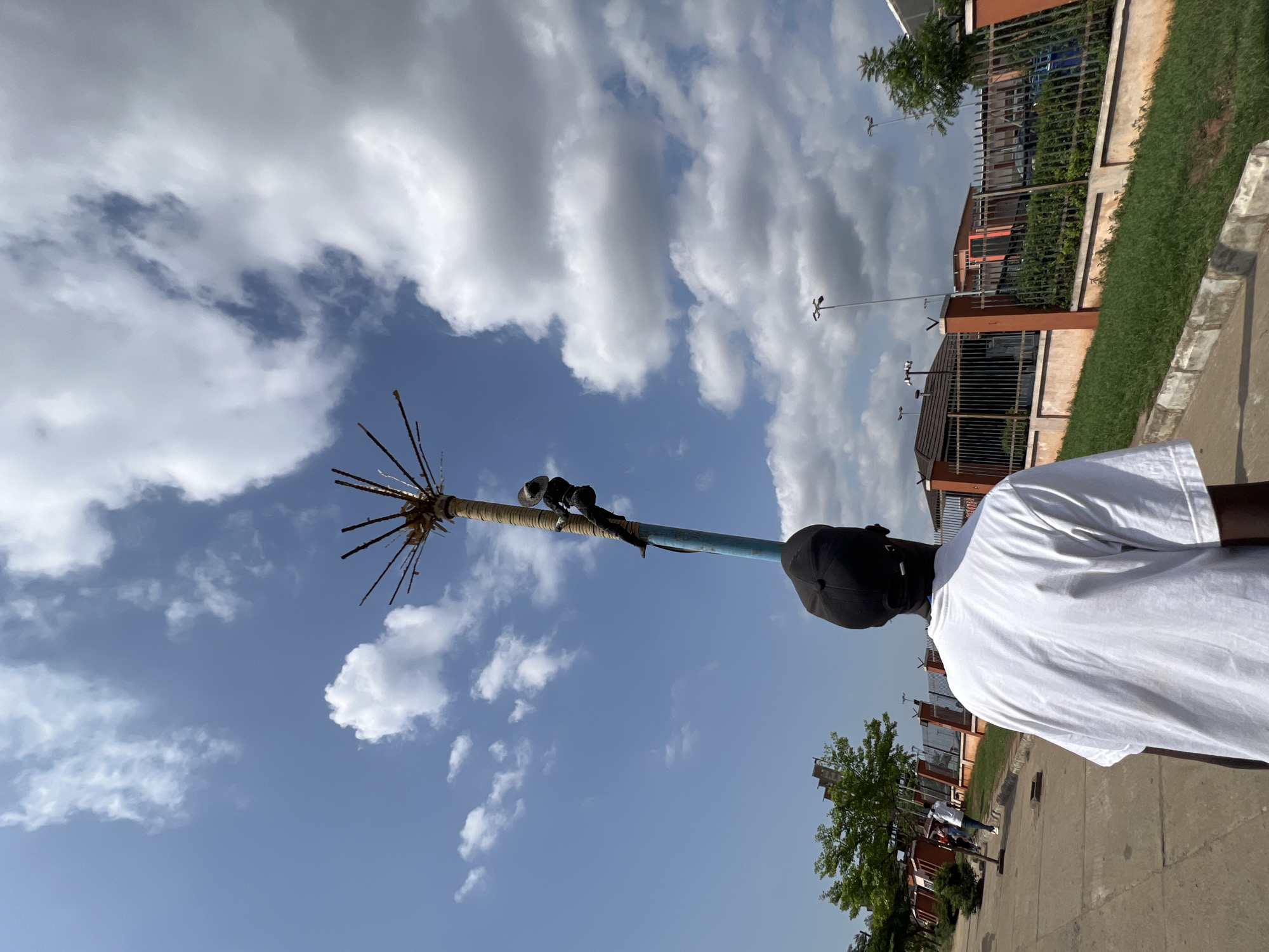 The image captures a bright daytime outdoor scene, likely on a street in Onitsha, Nigeria. The sky is a vibrant blue with scattered white cumulus clouds, suggesting fair weather.
In the foreground, positioned on the right side of the frame, is the back of a person wearing a plain white t-shirt and a black baseball cap. They are facing away from the camera, looking towards the left side of the scene.
A distinctive, tall, and slender pole stands prominently towards the center-left. Its lower section is painted a light blue, while the upper part is wrapped densely with what appears to be a light-colored rope or fabric. At the very top, numerous sharp, pointed sticks or rods radiate outwards, creating a unique, spiky, crown-like or starburst effect. A small, dark, cylindrical object, possibly a camera, sensor, or antenna, is attached to the wrapped section of the pole.
In the background, beyond the pole and the person, are several buildings with orange or reddish-brown walls and dark metal fences, typical of residential or commercial structures. Green grass borders the paved surface of what appears to be a street or sidewalk. Further down the street, several modern streetlights with multiple arms are visible. The overall scene depicts a typical urban or suburban setting under sunny conditions.