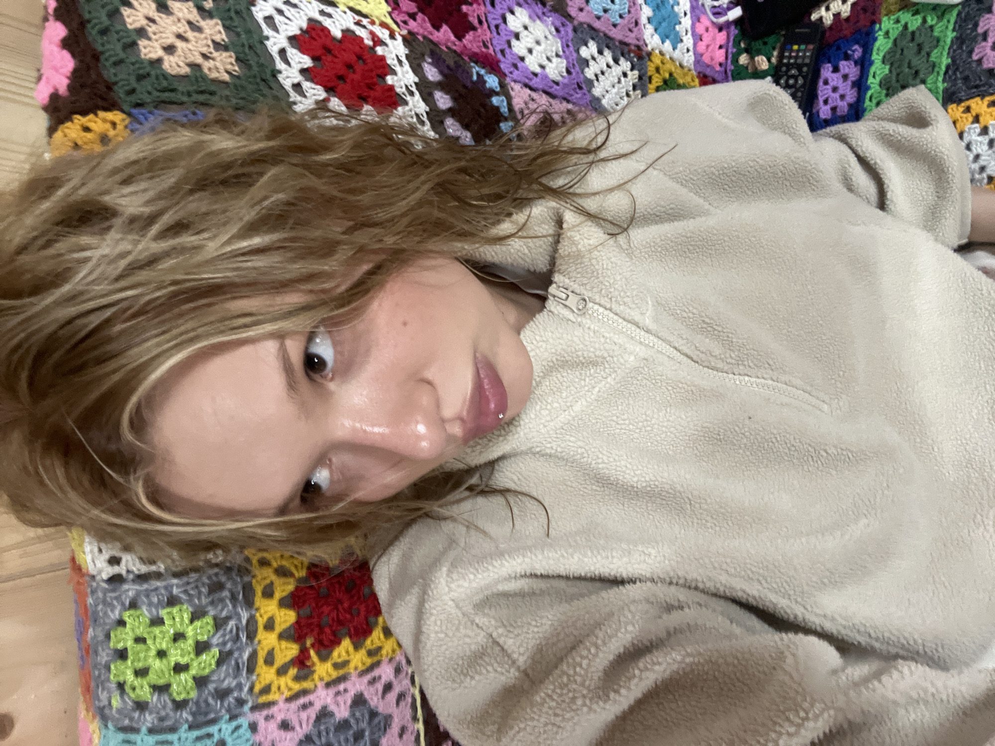 The image depicts a person with light brown, wavy hair and fair skin, wearing a beige fleece jacket with a zipper. The person is lying on a colorful crocheted blanket with various square patterns. A remote control is visible in the background. The location is unspecified but assumed to be Chaikovsky, Russia.