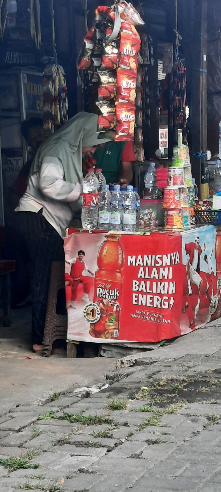 The image depicts a street-side stall in East Jakarta, Indonesia, likely operating in a neighborhood setting. In the foreground, a table displays a variety of beverages, including bottled water and a prominent banner advertising "Teh Pucuk Harum." The banner features a large image of the tea bottle, accompanied by text in Indonesian that translates to "Sweetness, Natural, Revitalize Energy" and "Without preservatives, Without artificial sweeteners." Two individuals are partially visible behind the table. One, wearing a light-colored hijab and patterned pants, is attending to the items on the table. Another person, wearing a green shirt, is seen behind the first individual. The stall is adorned with hanging snacks and other merchandise, typical of a small local convenience store. The pavement in front of the stall is made of interlocking gray pavers with patches of grass growing in the cracks. The overall scene suggests a typical day of commerce in a local Indonesian community.