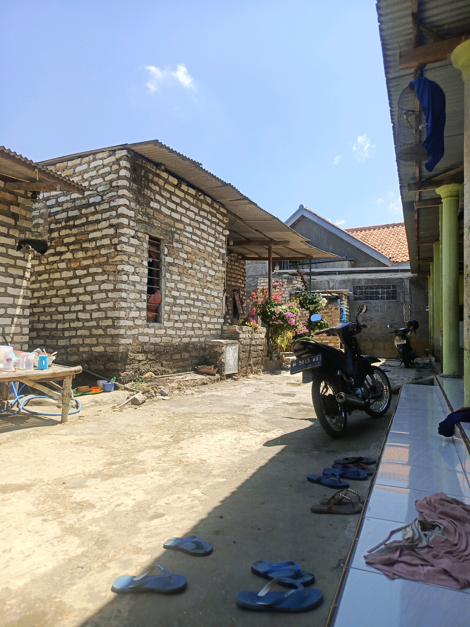 The image captures an outdoor courtyard, likely in a residential area of Bugih, Indonesia. The scene is characterized by a predominantly sunny day with a clear blue sky. In the foreground, a concrete ground is visible, scattered with several pairs of blue flip-flops and a brown pair. To the right, a tiled walkway leads into an open-sided structure supported by thick green pillars, with a pile of folded clothing on the tiles.
Dominating the middle ground is a black motorcycle parked on the concrete. Behind the motorcycle, another motorcycle is partially visible. Further back, a weathered brick building with a corrugated metal roof stands prominently. A window with bars on this building reveals a hint of orange fabric inside. To the left of the brick building, a simple bamboo table holds several plastic containers, a blue hose, and other miscellaneous items. Flower bushes with pink and white blooms are positioned near the brick structure.
In the background, more buildings with red-tiled roofs are visible, suggesting a densely populated area. A birdcage hangs from the eaves of one of the structures, covered partially by a blue cloth. The overall impression is one of a simple, lived-in environment, typical of many Indonesian villages or towns. There are no people visible in the image.