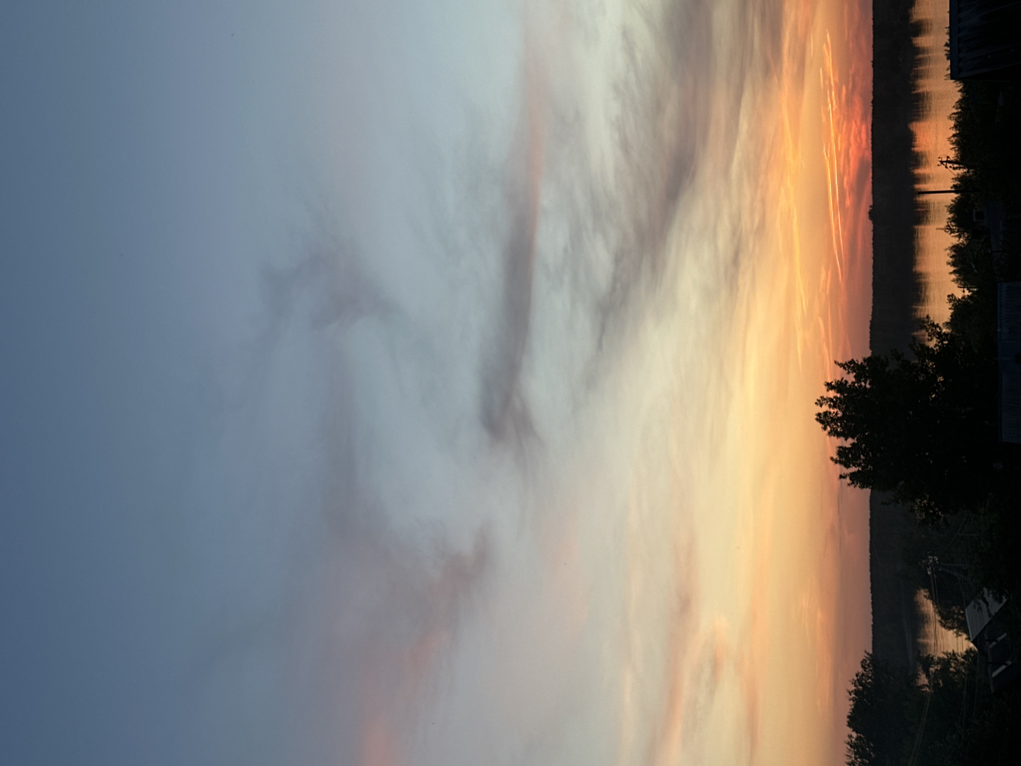 The image depicts a sunset scene, likely captured near a body of water, which is suggested to be in or near Chaikovsky, Russia, by the query. The sky transitions from a deep blue on the left to an orange-red hue towards the right, indicating the setting sun. Clouds are present, partially obscuring the sun and scattering its light. The horizon features a dark silhouette of trees and possibly some structures, suggesting a shoreline or a wooded area bordering the water. The water reflects some of the colors from the sky, creating a mirroring effect. The image is rotated counter-clockwise, which places the sky on the left and the landscape on the right. No people are visible in the scene.