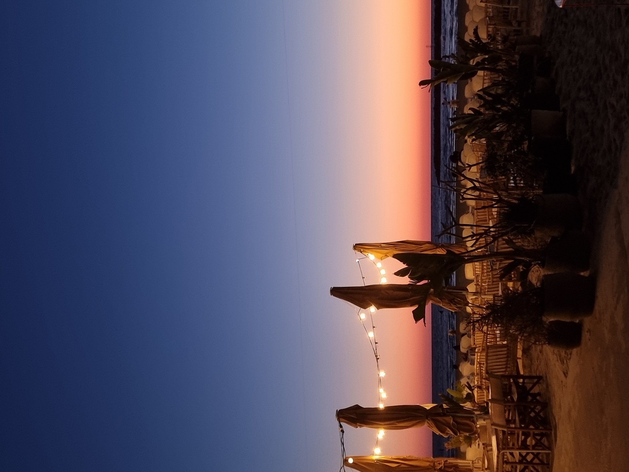 The image shows an outdoor restaurant setting at sunset, likely in Tel Aviv, Israel, given its coastal location and ambiance. The sky transitions from deep blue to orange, indicating the time of day. The restaurant is on a beach, evident from the sand along the right edge. Tables and chairs are visible, many under straw umbrellas adorned with string lights, which are illuminated. Planters with tropical plants are strategically placed. The sea is barely visible in the background, and some people are seated at the tables.