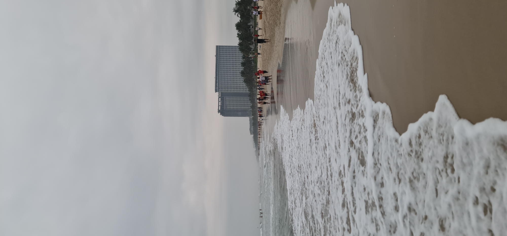 The image captures a panoramic view of a beach in Đà Nẵng, Vietnam, under an overcast sky. The scene is oriented vertically, showing the expansive beach stretching into the distance.
On the right side of the frame, frothy white ocean waves actively crash and recede along the light brown sandy shore. The water extends towards the horizon, where it meets the dull gray sky. The left side of the image features the wide, flat beach.
Numerous people are visible along the beachfront. Many individuals are scattered across the sand, walking, standing, or congregating in small groups. A notable cluster of people, many dressed in red shirts, is seen closer to the background buildings, suggesting a group event or activity. Further out in the shallow water, several people are wading or swimming.
In the background, a row of tall, modern multi-story buildings, consistent with hotels or residential complexes, lines the coastline. These structures rise above a continuous band of green palm trees that separate the buildings from the sandy beach.
The sky is uniformly overcast, casting a soft, muted light over the entire scene, indicating either an early morning, late afternoon, or simply a cloudy day. There is no visible text in the image. The overall setting is a developed and popular urban beach destination.