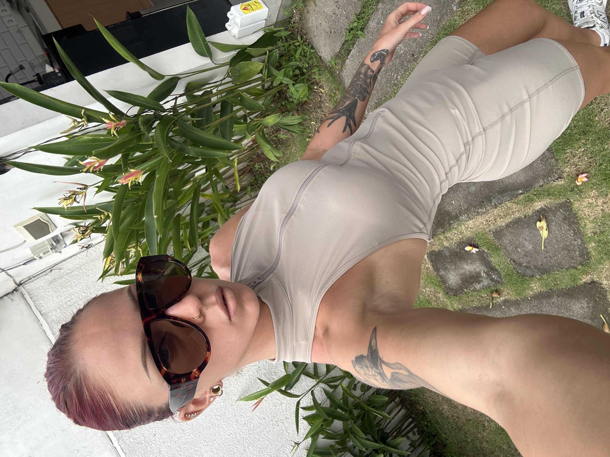 The photograph shows a woman wearing a tan-colored bodysuit and sunglasses. She is standing outdoors in a location with a garden or landscaping, with green plants visible around her. The background includes a white building and a stone pathway. The photograph was taken in the city of North Kuta, Indonesia.