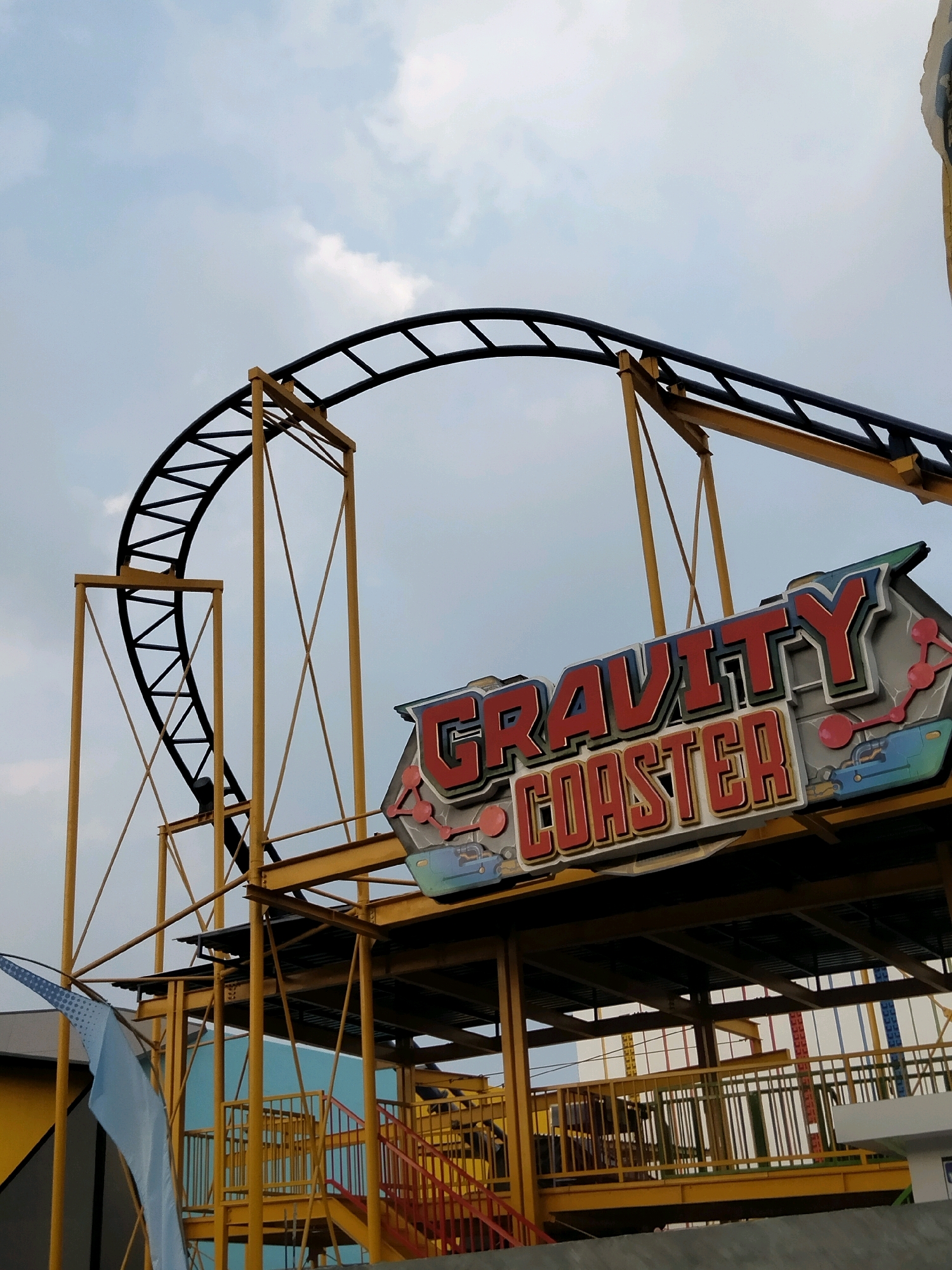 The image shows a close-up view of the "Gravity Coaster" attraction at Museum Angkut in Batu, Indonesia. The coaster track, made of black metal and supported by yellow steel structures, curves upwards against a cloudy sky. A prominent sign with red and white lettering that reads "GRAVITY COASTER" is visible on the right side of the frame, adorned with colorful graphics. Below the track and sign, yellow platforms and railings are visible, suggesting the boarding area or a section of the ride's structure. The overall scene depicts an amusement park setting.