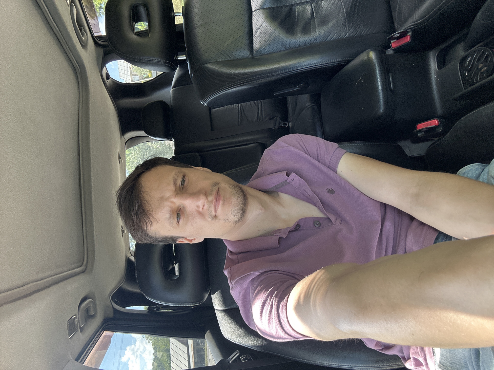 A man with short brown hair and a light beard is seated in the passenger seat of a car, taking a selfie. He is wearing a short-sleeved purple polo shirt and blue jeans. The interior of the car is visible, featuring black leather seats, a center console with cup holders, and a sunroof. Sunlight streams into the car, casting shadows on the man's arm and the interior. Outside the car, trees and a building are visible, suggesting an outdoor setting. The scene appears to be in Makiv, Ukraine.