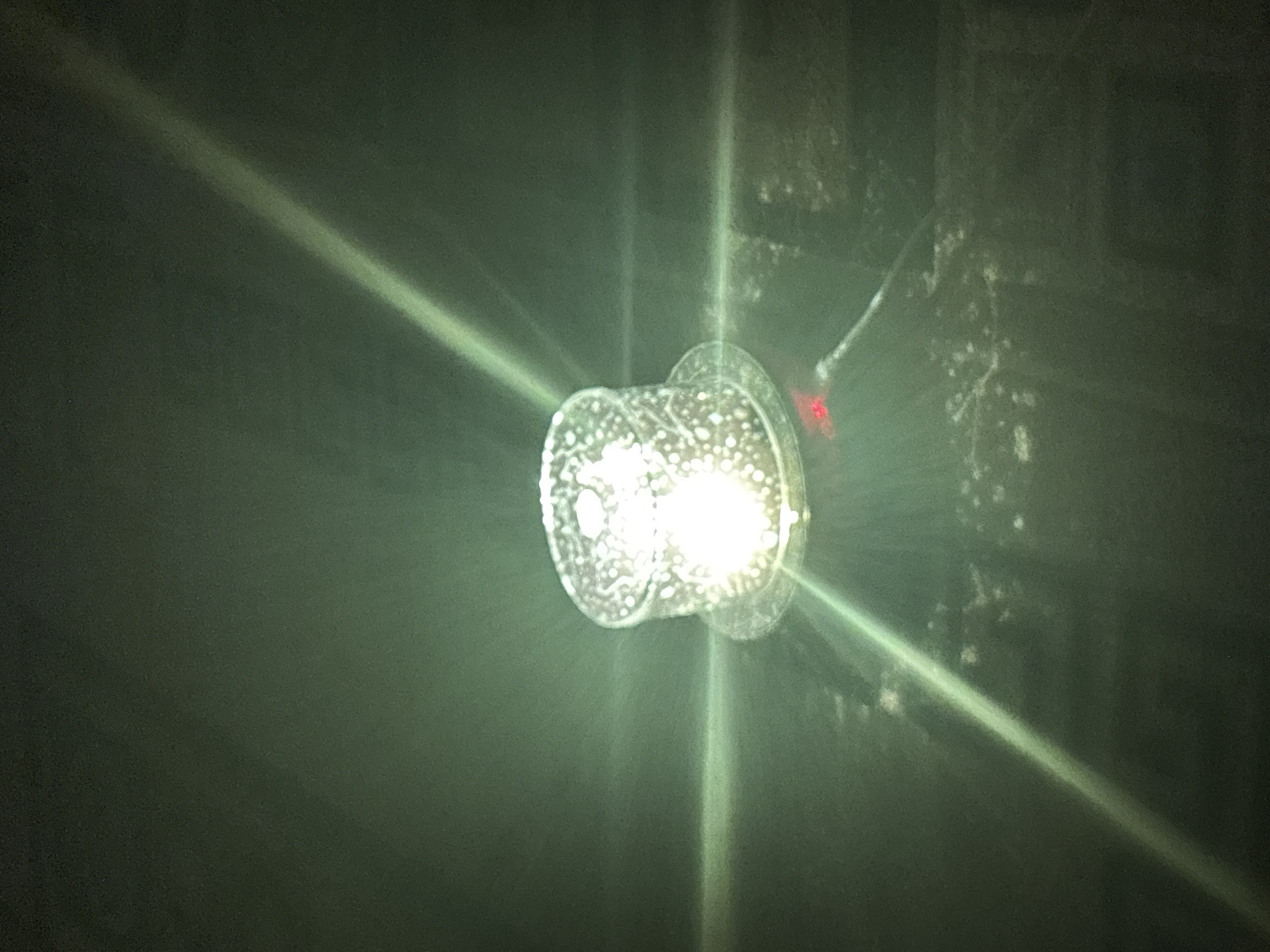 An illuminated, cylindrical object with a textured surface sits on a dark, reflective surface. Bright light emanates from within the object, creating strong lens flares that extend horizontally and diagonally across the frame. The surrounding environment is dark and appears to have a textured or uneven surface with some scattered light-colored specks. There are no people or identifiable landmarks present in the image. The scene does not provide any specific clues to its location within the city of Kryvyi Rih, Ukraine.