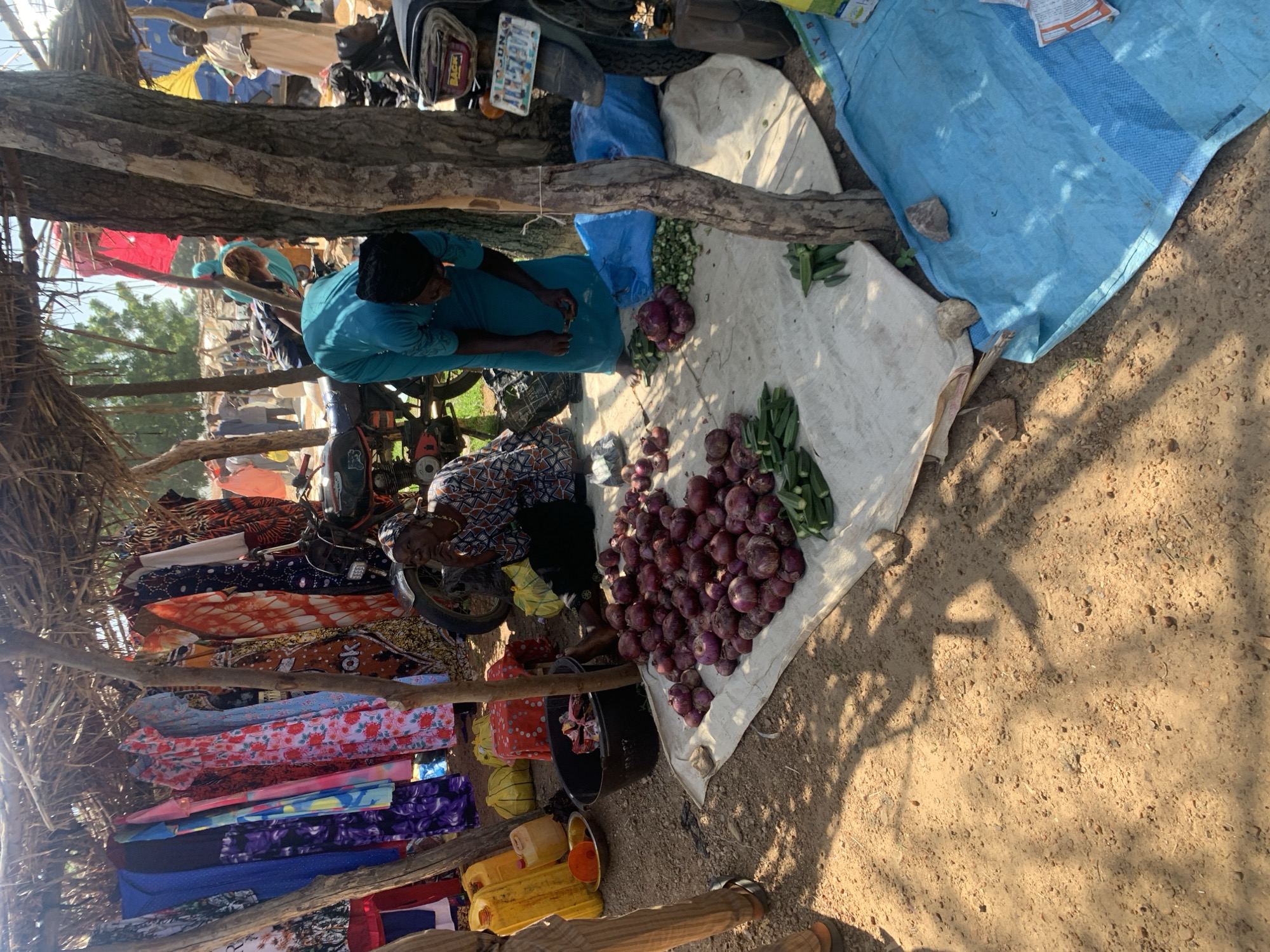 This image depicts an outdoor market scene in Boi A, Nigeria, characterized by a makeshift stall constructed with natural materials like straw and wood. The stall displays a variety of colorful clothing items, predominantly dresses, hanging from a rail. In the foreground, a woman is seated on the ground next to a pile of red onions and okra arranged on a white cloth. Another woman, wearing a blue garment, is standing and appears to be sorting vegetables. A motorcycle is partially visible, suggesting it is a mode of transport or a vendor's item. The ground is sandy and uneven, with shadows from overhead structures and trees indicating a sunny day. Other market activities and people are visible in the background, suggesting a bustling environment. The overall impression is that of a typical local market in a rural or semi-urban setting in Nigeria.