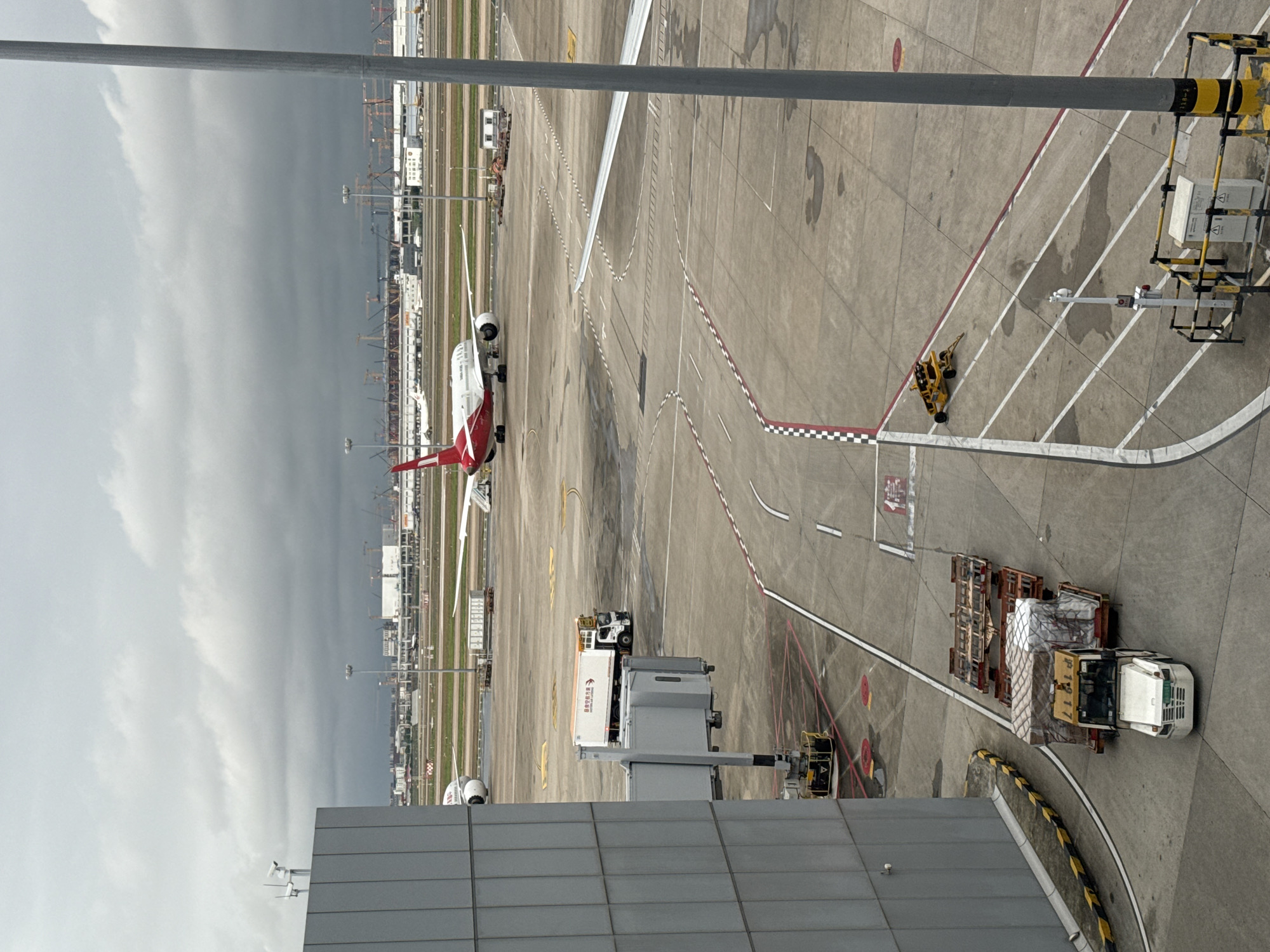 The image depicts an airport tarmac in Pudong, China, under a cloudy sky. A large passenger airplane with red and white livery is parked on the runway in the background. In the foreground, a jet bridge extends from a modern terminal building. Several service vehicles are visible on the tarmac, including a white tow tractor pulling a cart laden with cargo, a refrigerated truck, and a yellow baggage tug. Construction cranes are present in the distance, suggesting ongoing development at the airport. The ground is paved concrete, marked with various lines and symbols indicating taxiways and runways. The scene is typical of an operational airport environment.