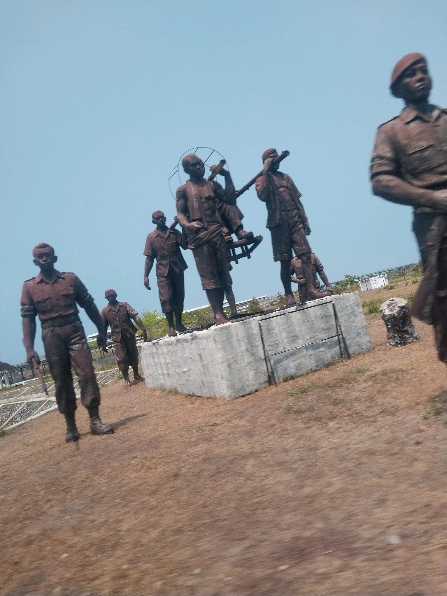 This image shows a collection of bronze statues depicting soldiers. The statues are in various poses, with some standing and others carrying objects. They appear to be placed outdoors in a dry, grassy area. In the background, there are some low buildings and a clear blue sky. The statues are dark brown in color, and some of them are worn or damaged. The scene is likely a memorial or monument, given the subject matter. The location is identified as Palihan, Indonesia.