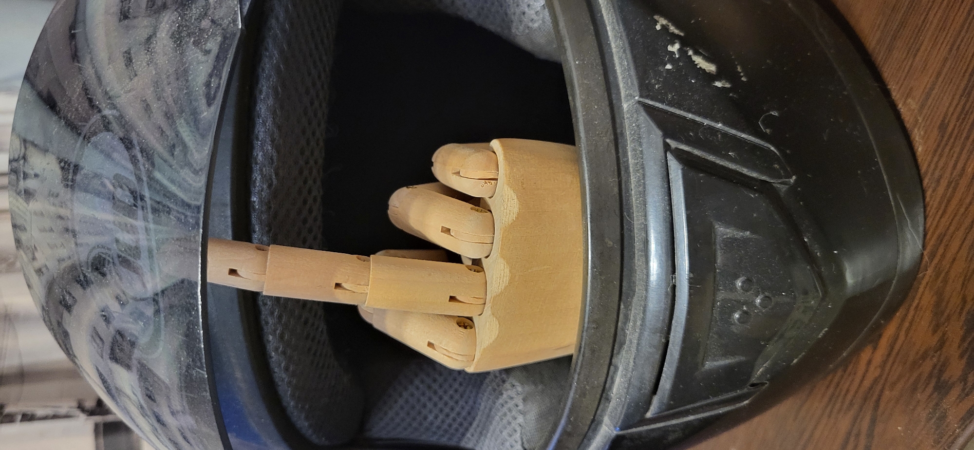 A wooden mannequin hand is positioned inside a black motorcycle helmet, with the middle finger extended upwards. The helmet is black with a swirling grey and white pattern on the exterior, and it appears dusty. The interior of the helmet is lined with grey fabric. The helmet rests on a dark wooden surface, possibly a table. The image is a close-up shot, focusing on the helmet and the hand. The background is out of focus and appears to be a wall with some vertical lines. There is no indication of the city of Almaty, Kazakhstan, or any urban environment.