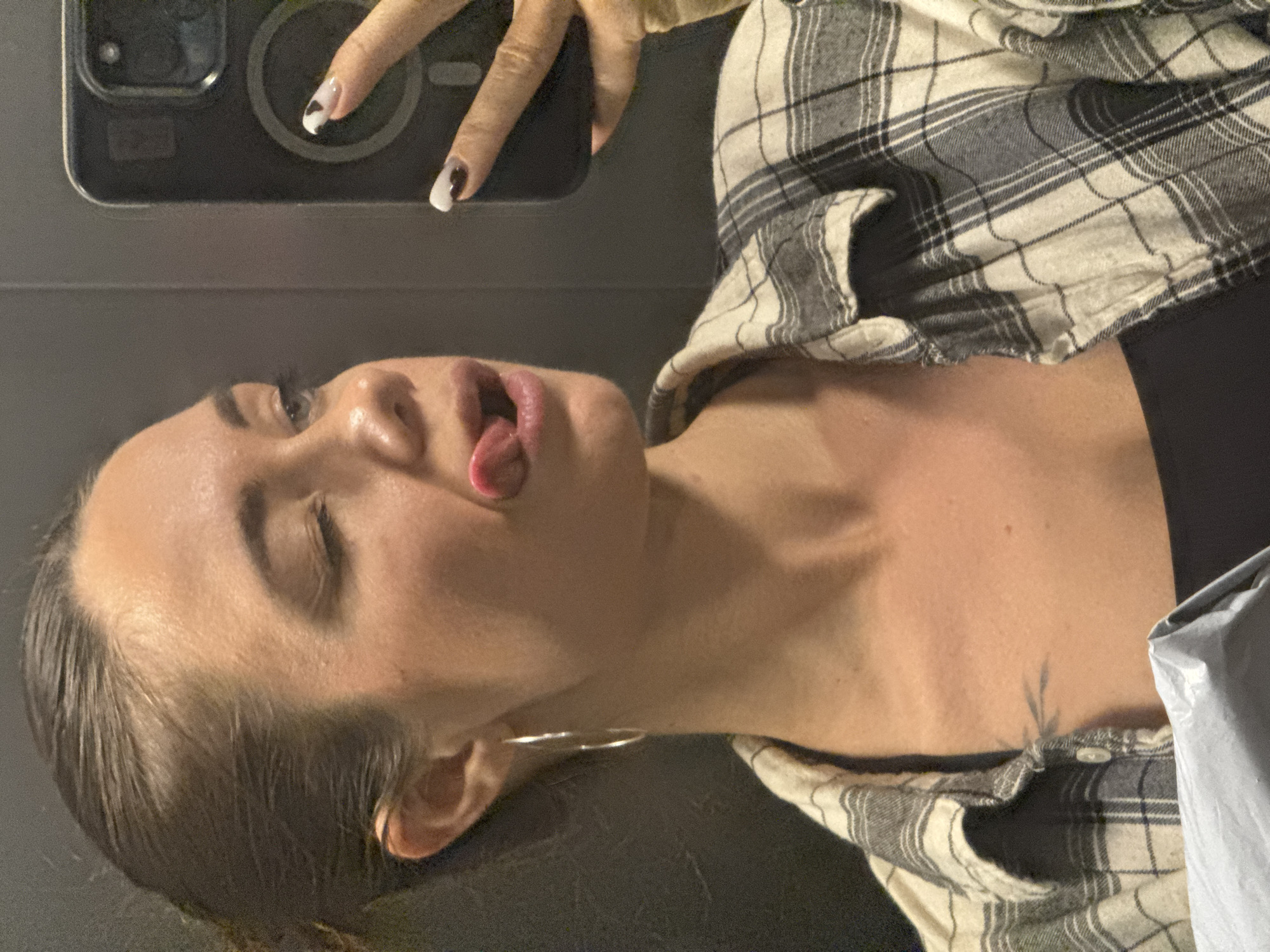A woman is taking a selfie with her phone. She is wearing a plaid shirt and has a small tattoo on her chest. Her tongue is sticking out. The location is not discernible from the image.